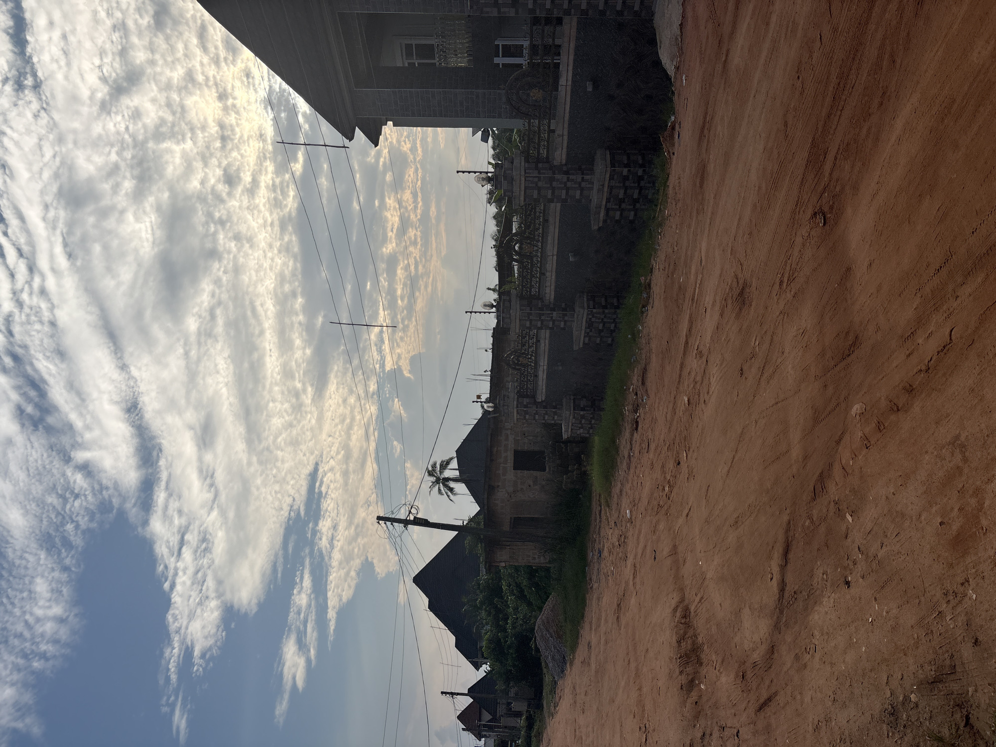The image depicts a dirt road in a suburban area, likely Obosi, Nigeria, with several houses and a distinct cloudy sky. The foreground is dominated by the reddish-brown dirt road, showing tire tracks and some scattered debris. To the left, a line of houses with dark, pitched roofs is visible, nestled behind lush green vegetation. One house in the mid-ground appears to be under construction or in a state of disrepair, with exposed brickwork. A prominent feature in the mid-ground is a tall utility pole with numerous wires running from it.
On the right side of the image, a more substantial, modern house stands out. It has dark, textured walls, multiple windows, and a prominent balcony. This house is enclosed by a decorative stone and metal fence. The fence is elaborate, featuring stone pillars and intricate metalwork with scroll designs.
The sky above is a striking feature, filled with scattered, textured clouds, some of which are illuminated by the sun, creating a bright, almost hazy effect. The overall impression is one of a developing residential area, possibly on the outskirts of a Nigerian city, with a mix of older and newer construction styles.