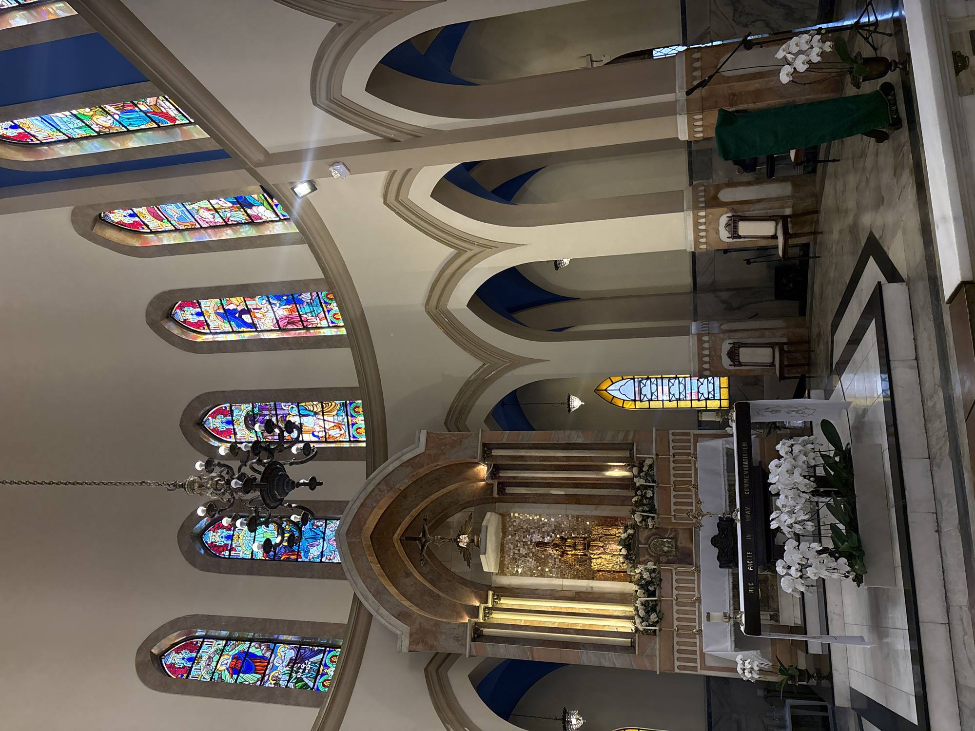 The image shows the interior of a church. The church has high ceilings with arched windows filled with stained glass. A large chandelier hangs from the ceiling. In the center of the image, there is an altar with a crucifix and a statue. White flowers are arranged around the altar. Pews are visible in the foreground. The architecture is ornate and religious in nature. This appears to be the interior of the Igreja Presbiteriana Independente de São Paulo.