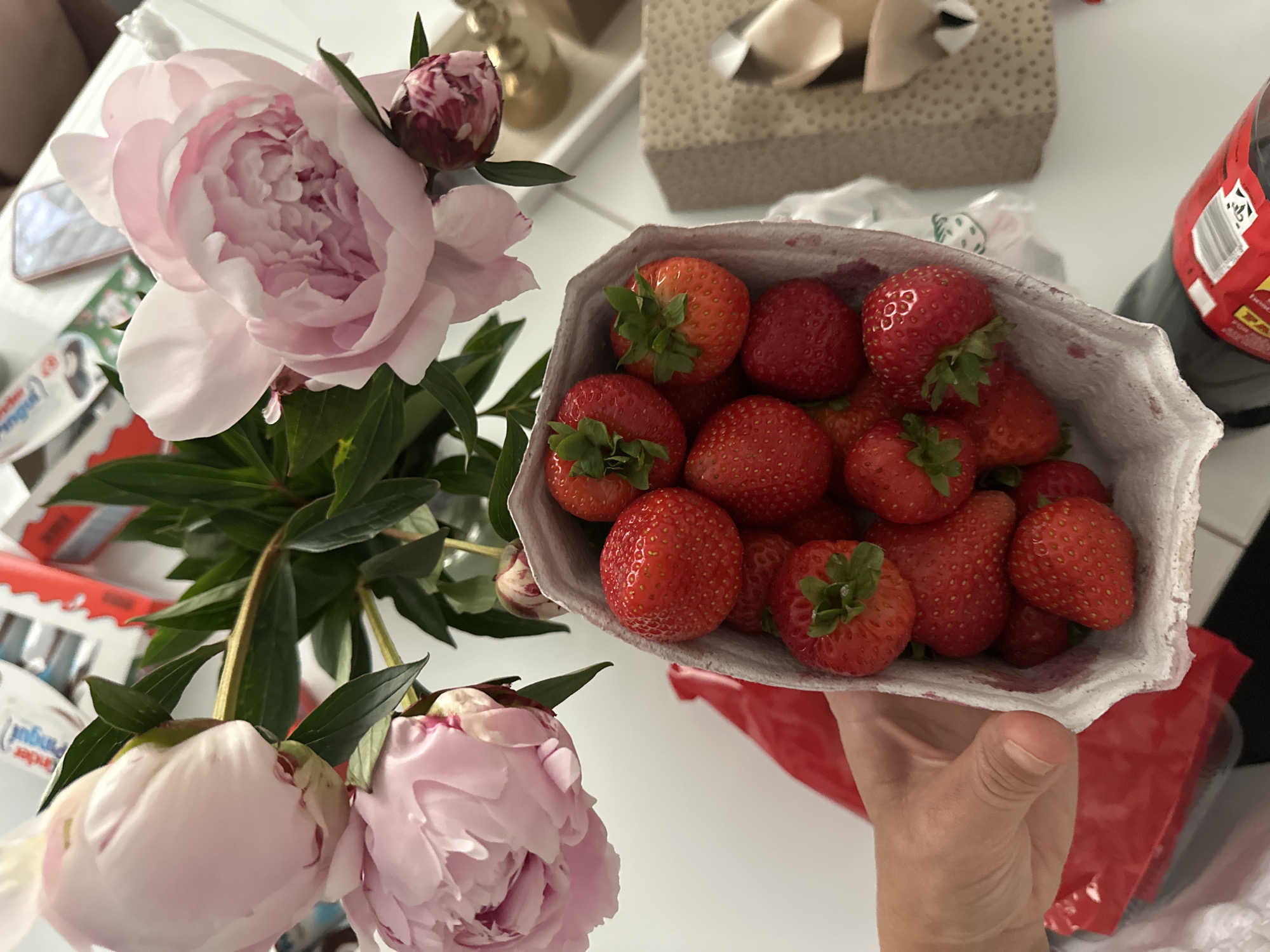 The image depicts a close-up overhead view of a hand holding a white paper carton filled with ripe strawberries. The carton is angled, revealing multiple bright red strawberries with green stems. In the background, a bouquet of pink peonies with green leaves is arranged. To the right of the peonies, a light-colored surface with what appears to be a gift-wrapped box is visible, along with a metallic object. Further back, a mobile phone with a light pink case rests on the same surface. In the upper background, partially obscured, are packages with red and white branding, possibly candy or food items. The overall setting appears to be indoors, possibly a home environment in Kutenholz, Germany, given the context provided.