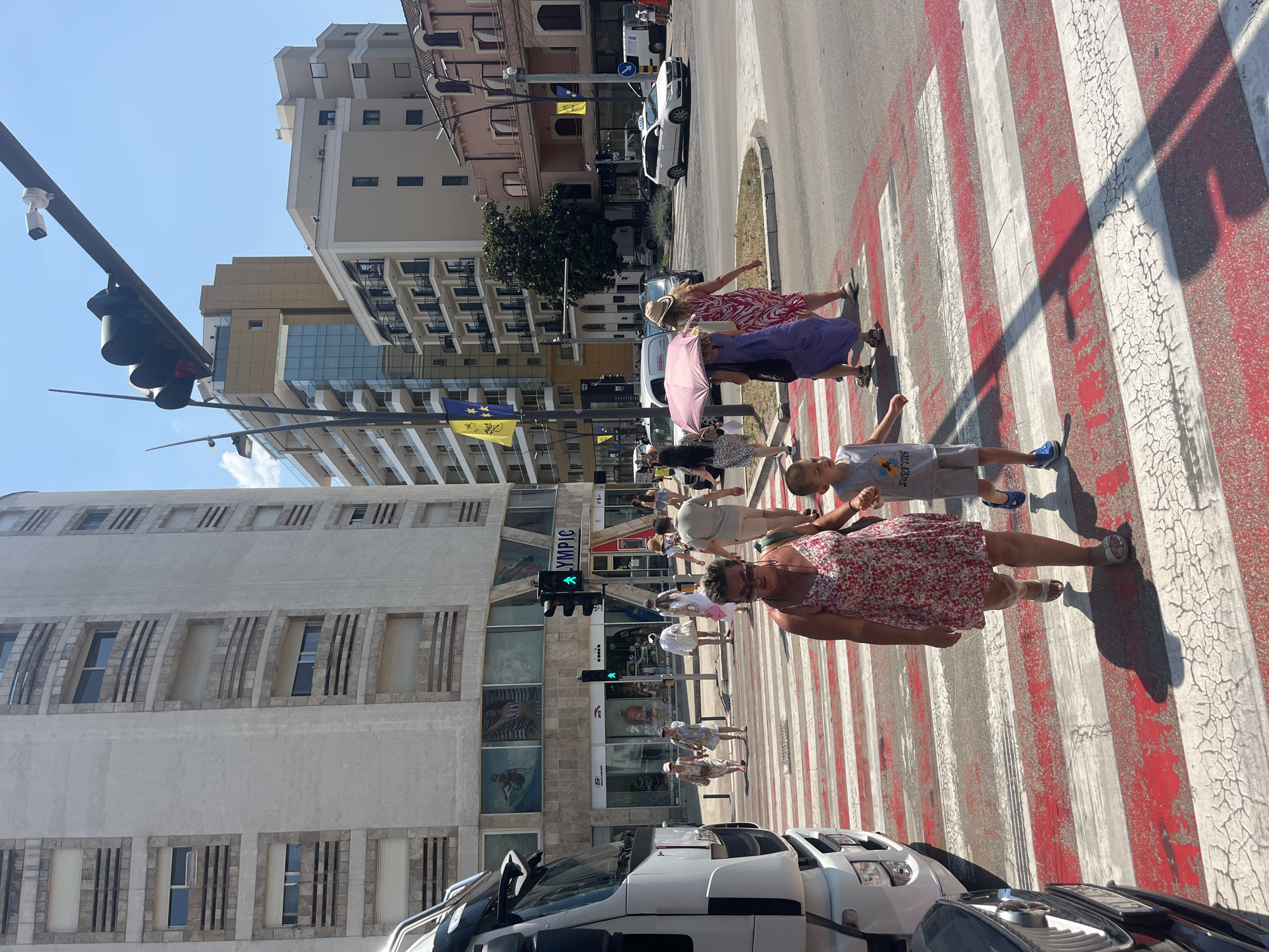 A bright, sunny day in an urban environment features a group of people crossing a street at a pedestrian crossing. In the foreground, a woman in a floral dress and a young boy in a grey t-shirt and shorts are walking across the red and white striped zebra crossing. The woman is wearing sunglasses and the boy is looking to his left. Behind them, other pedestrians are also crossing, including a woman in a purple dress and another in a pink and white patterned dress, one of whom is holding a pink umbrella. A large white truck is parked on the left side of the image, partially obscuring the view of a building. The building on the left has multiple floors with rows of windows and appears to be a commercial establishment with signage visible, including "OLYMPIC". Other buildings, some modern with balconies and glass facades, and others older with arched windows, are visible in the background. Traffic lights are visible, with a green walking signal illuminated. The sky is clear and blue. The scene appears to be a bustling city street on a warm day.