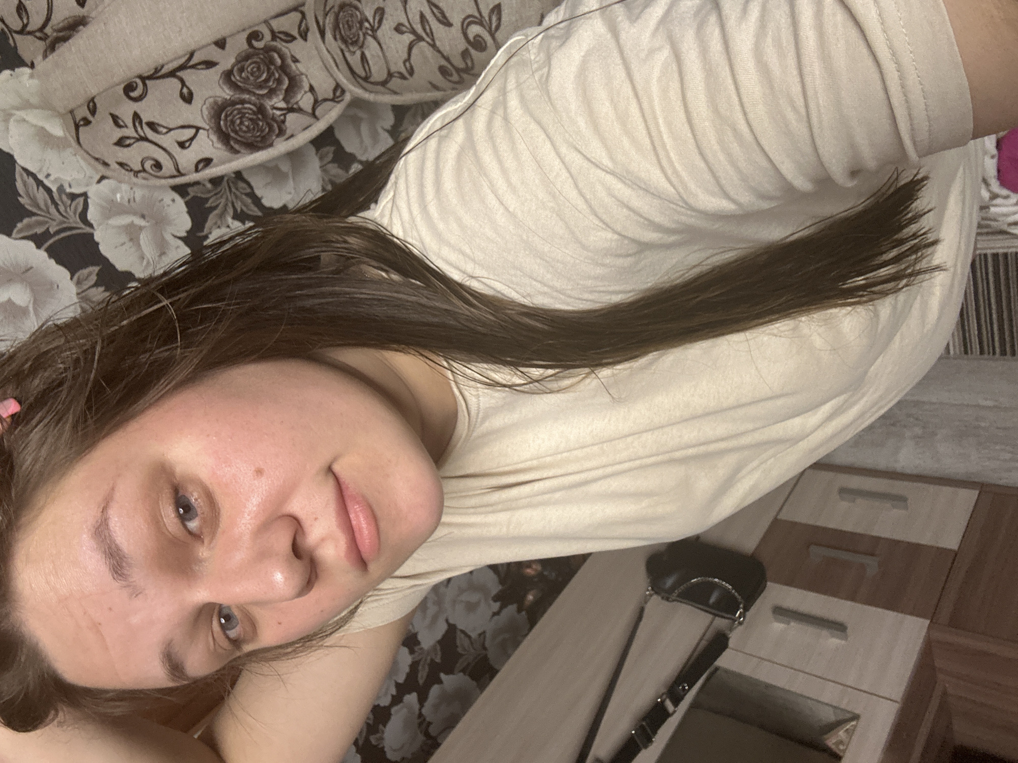 A woman is lying on a floral patterned couch in what appears to be a home interior. She is wearing a light-colored t-shirt. Her hair is long and brown. A black purse hangs off a nearby wooden dresser. There is no indication of the location being in the city of Ольховка, Russia.