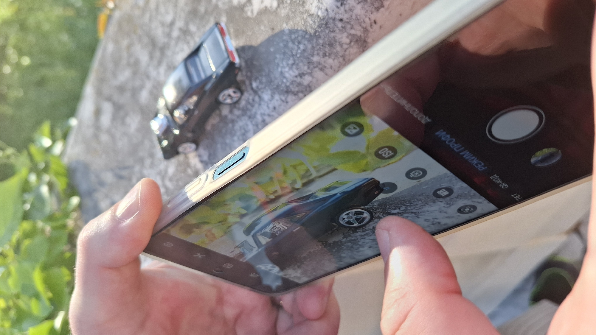 A person is holding a smartphone in their hands, taking a picture of a black toy car on a concrete surface. The smartphone screen displays the image of the car that the camera is capturing. The background consists of foliage and blurred elements. The location appears to be outdoors, possibly in a garden or on a patio.