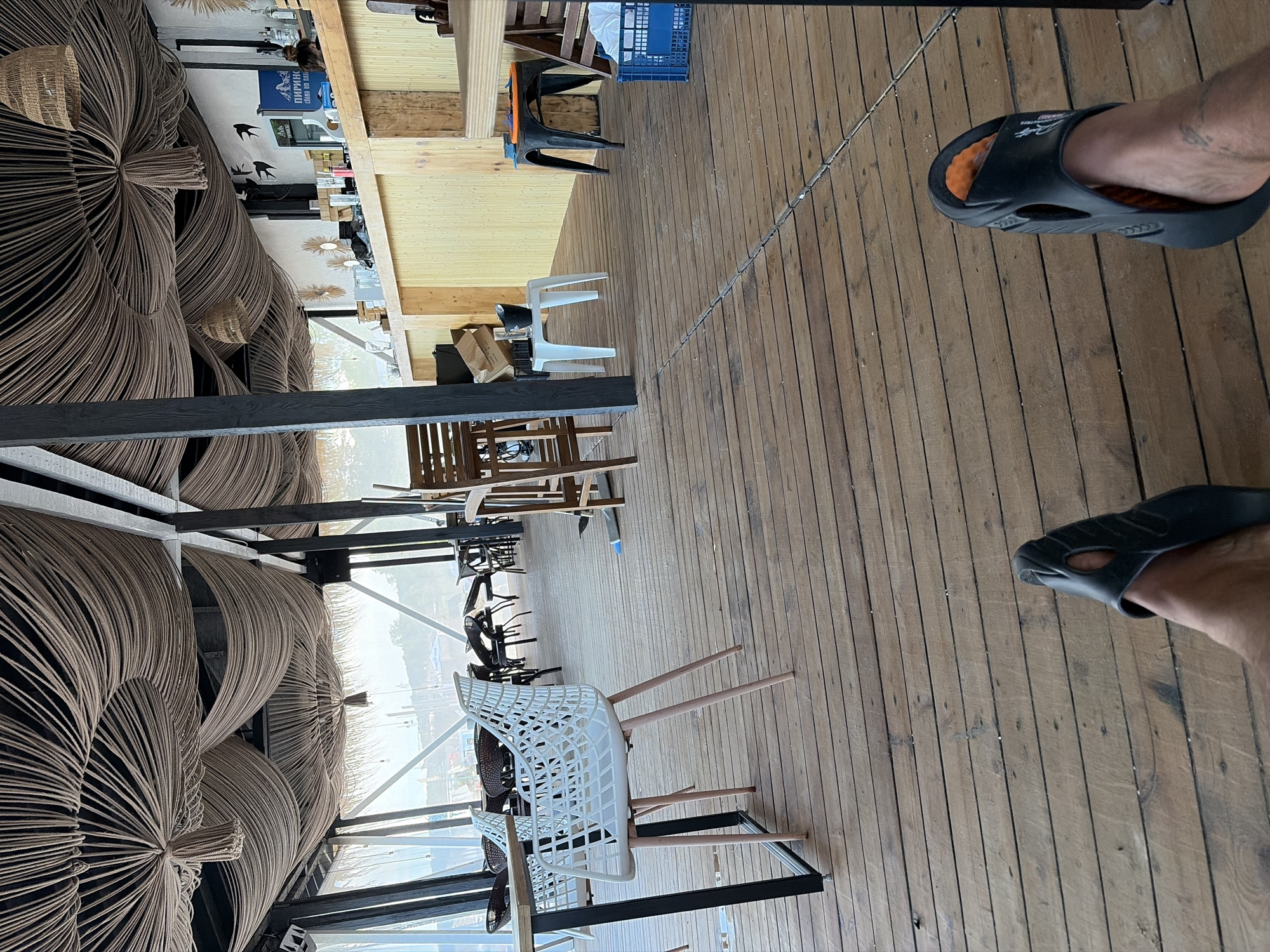This image depicts an outdoor seating area with a wooden floor and a ceiling covered in decorative rope. The space appears to be part of a restaurant or bar, with tables and chairs visible. In the background, a bar counter with bottles and a refrigerator can be seen. The setting is open-air, with a view of a hazy outdoor landscape. The foreground shows a person's legs and feet wearing black slides, resting on the wooden floor. The overall atmosphere suggests a relaxed, casual environment, possibly at a beachside establishment in Sozopol, Bulgaria, given the context provided. The lighting indicates it is daytime, with natural light filtering into the covered area.