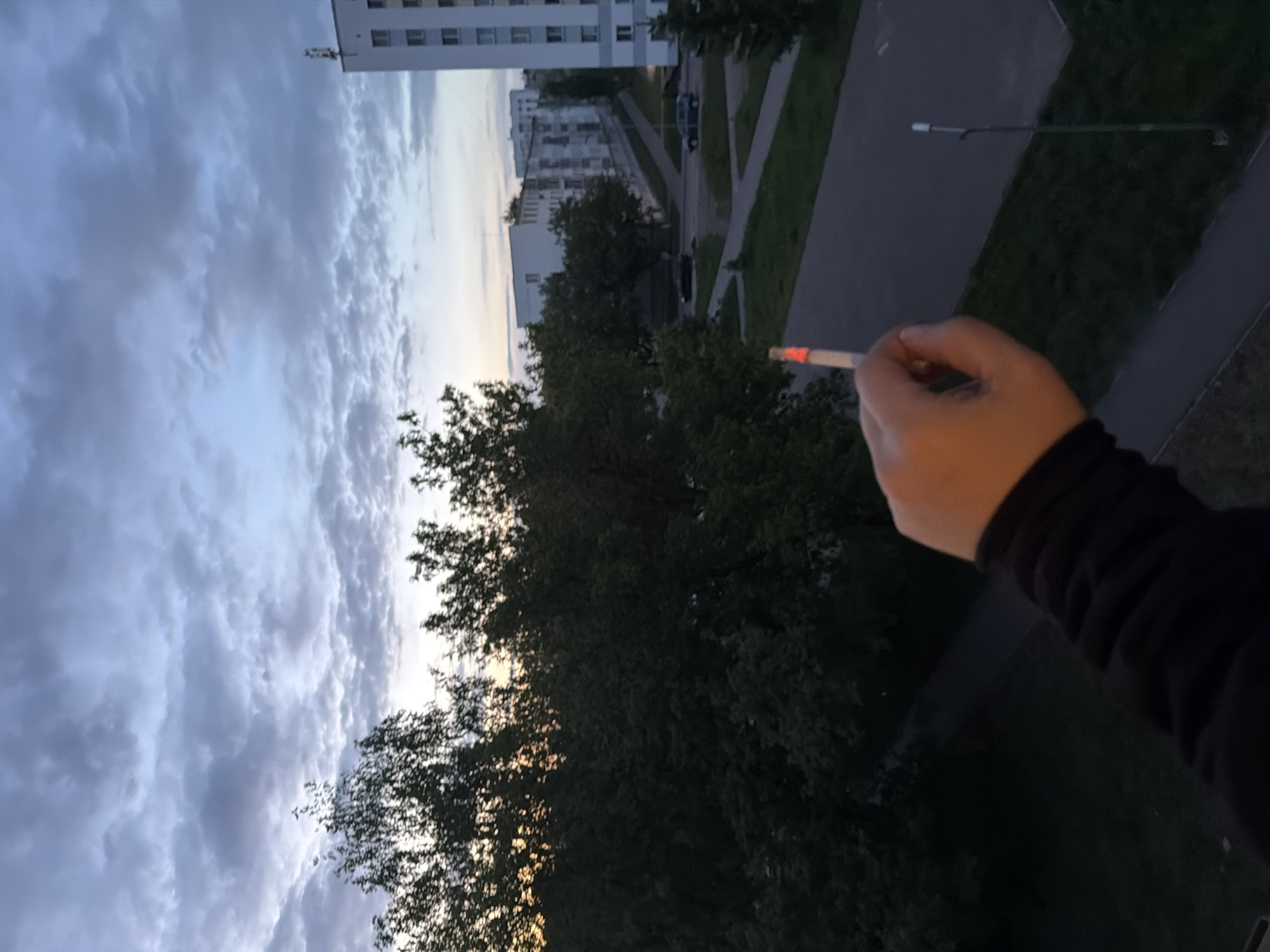 A photograph shows a hand holding a lit cigarette against a backdrop of trees and a building, partially obscured by the trees. The sky is filled with clouds. A sidewalk is visible in the upper right. The photograph was taken in Naberezhnye Chelny, Russia.
