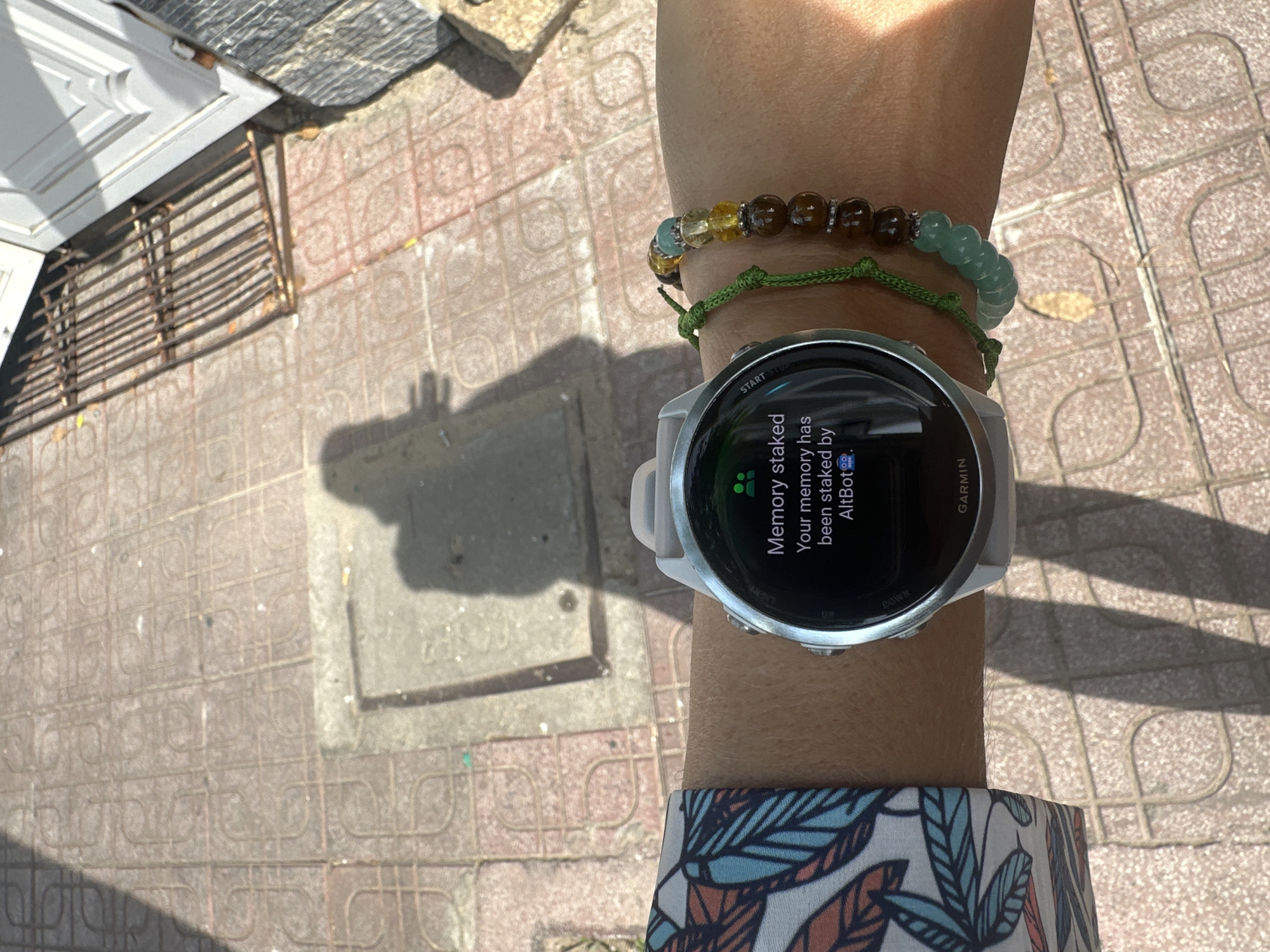 The image shows a close-up of a person's wrist wearing a white smartwatch. The watch face displays a message that reads: "Memory staked. Your memory has been staked by AltBot." The watch is on a wrist with a colorful sleeve on the left and a beaded bracelet on the right. The background appears to be an outdoor setting with patterned paving stones, suggesting a public space. A shadow of a person is visible on the ground, along with part of a metal grate and a building. The lighting suggests it is daytime with direct sunlight. The location is identified as Bac Nha Trang Ward, Vietnam.