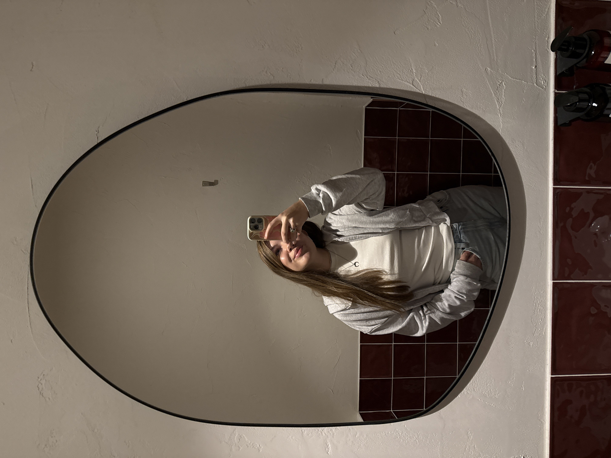 The photograph captures a mirror selfie taken in a bathroom with distinctive design elements, presumably in Belgrade, Serbia (though there's no definitive visual confirmation of the location).
A young woman is seen in the mirror's reflection, holding her phone up and appearing to take a picture. She is wearing a white t-shirt, a grey zip-up hoodie, and jeans. Her hair is long and light brown.
The mirror itself is oval-shaped with a black frame, mounted on a textured beige-colored wall. To the right of the mirror are dark red square tiles. A dispenser is visible on the tiled area. The composition of the mirror and wall suggests a modern interior design.