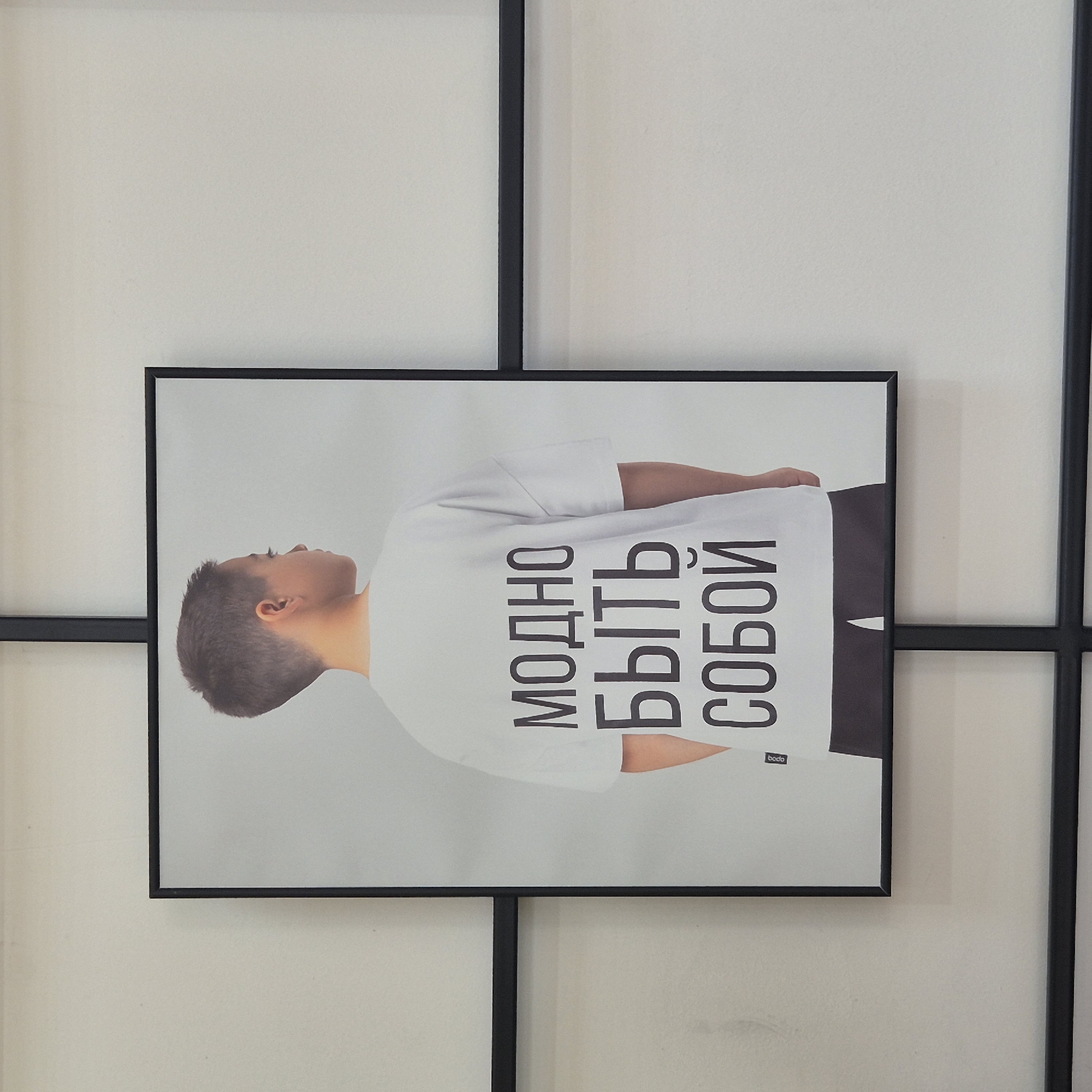 A framed picture hangs on a wall. The picture features a young boy from behind, wearing a white t-shirt with dark Russian text printed on it. The text reads "МОДНО БЫТЬ СОБОЙ," which translates to "It's fashionable to be yourself." The boy has short, dark hair and is looking to his right. The wall is a light, neutral color and has a grid pattern created by black frames or beams. The image is captured at an eye-level perspective, with the picture occupying the majority of the frame. The overall lighting appears to be soft and even. The location of this image is Kokshetau, Kazakhstan.