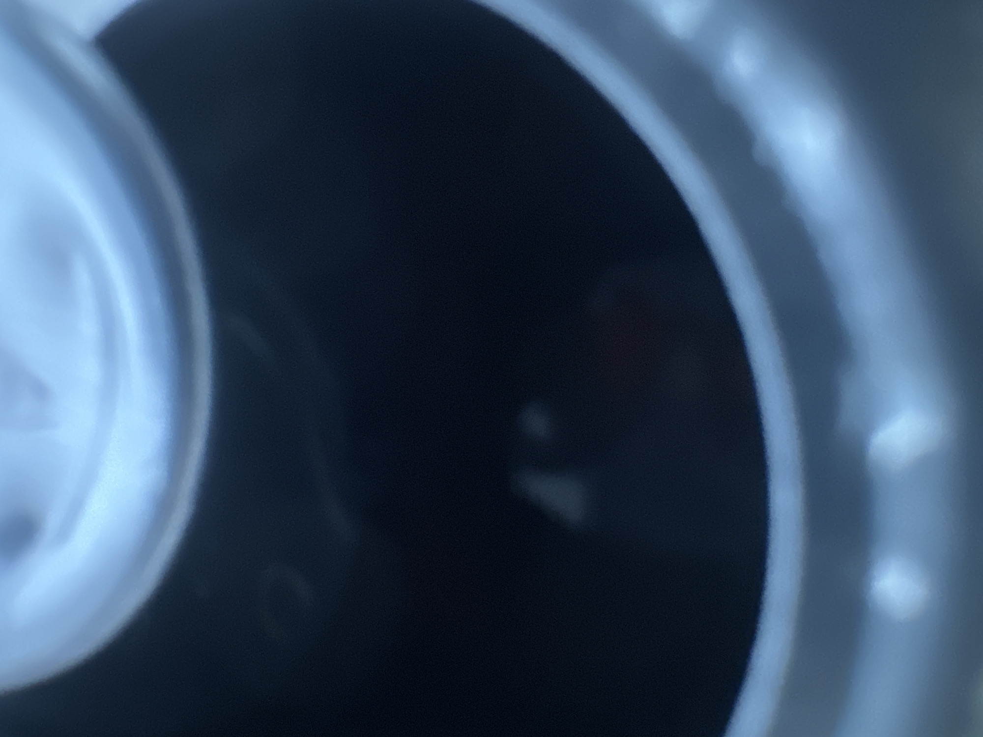 The image is an extreme close-up shot looking down into a dark, cylindrical object, possibly a can or a pipe. The rim of the object is visible at the top and bottom, appearing to be made of a light-colored material, likely metal, with some reflective qualities. The interior is predominantly dark, with some diffuse light sources creating subtle highlights and shadows. In the lower center of the image, there are two indistinct, blurry white or light gray shapes, possibly reflections or small objects within the container. The overall impression is one of looking into a confined, dark space from above. The location of this image in Belgrade, Serbia, cannot be determined from the visual information provided.