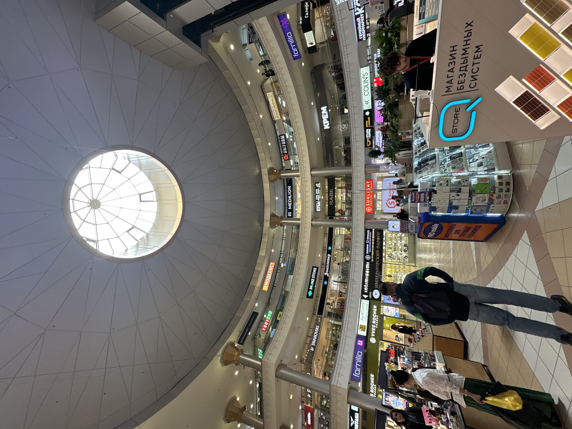 Here is a detailed description of the image:
**General Description:** The image captures an interior view of a shopping mall or large retail space, likely located in Saint Petersburg, Russia, given the Cyrillic text on store signs. The shot is taken from a slightly angled perspective, looking upwards to showcase the architecture and the overall layout of the mall.
**Architectural Details:** A massive dome-shaped ceiling dominates the top left portion of the image. The ceiling is segmented with a geometric pattern and has a large circular skylight at its center, allowing natural light to enter the space. The mall features multiple levels or floors, connected by open corridors and supported by large columns.
**Storefronts and Signage:** The image captures storefronts and various store signs in Cyrillic. Numerous store names are visible, including "Familia", "Swarovski", "Colin’s", "Yves Rocher", and "Sunlight". The signs are vertically oriented and layered along the corridors, which is a common feature of multi-level malls.
**People:** There are several people visible in the image:
* A man wearing a dark jacket and jeans is seen walking away from the camera.
* Various individuals are near storefronts.
* A shop assistant at a display counter.
**Objects and Displays:** In the foreground, there is a display counter for a "Store" that sells "Smokeless Systems". Mobile phone accessories are displayed on shelving.
**Lighting:** The mall is well-lit, primarily with artificial lighting supplemented by natural light from the skylight.
**Overall Impression:** The image conveys a sense of bustling activity in a modern, multi-level shopping mall. The architectural features and a variety of retail establishments create a dynamic and vibrant indoor environment.