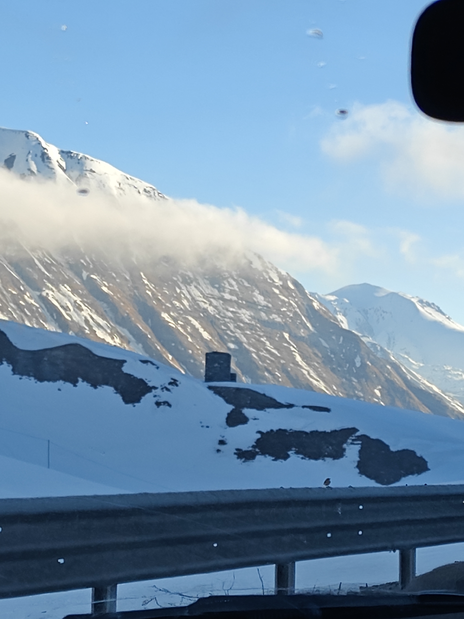 The image shows a landscape of snow-covered mountains under a partly cloudy sky. In the foreground, a metal guardrail is visible, with a small bird perched on it. A dark, square structure is centered on a snow-covered ridge. Behind the ridge, mountains rise steeply, partially obscured by clouds, with patches of snow. The image is likely taken from inside a vehicle due to reflections on what could be the car window. Based on the prompt information, the location is in the city of Ganisi, Georgia.