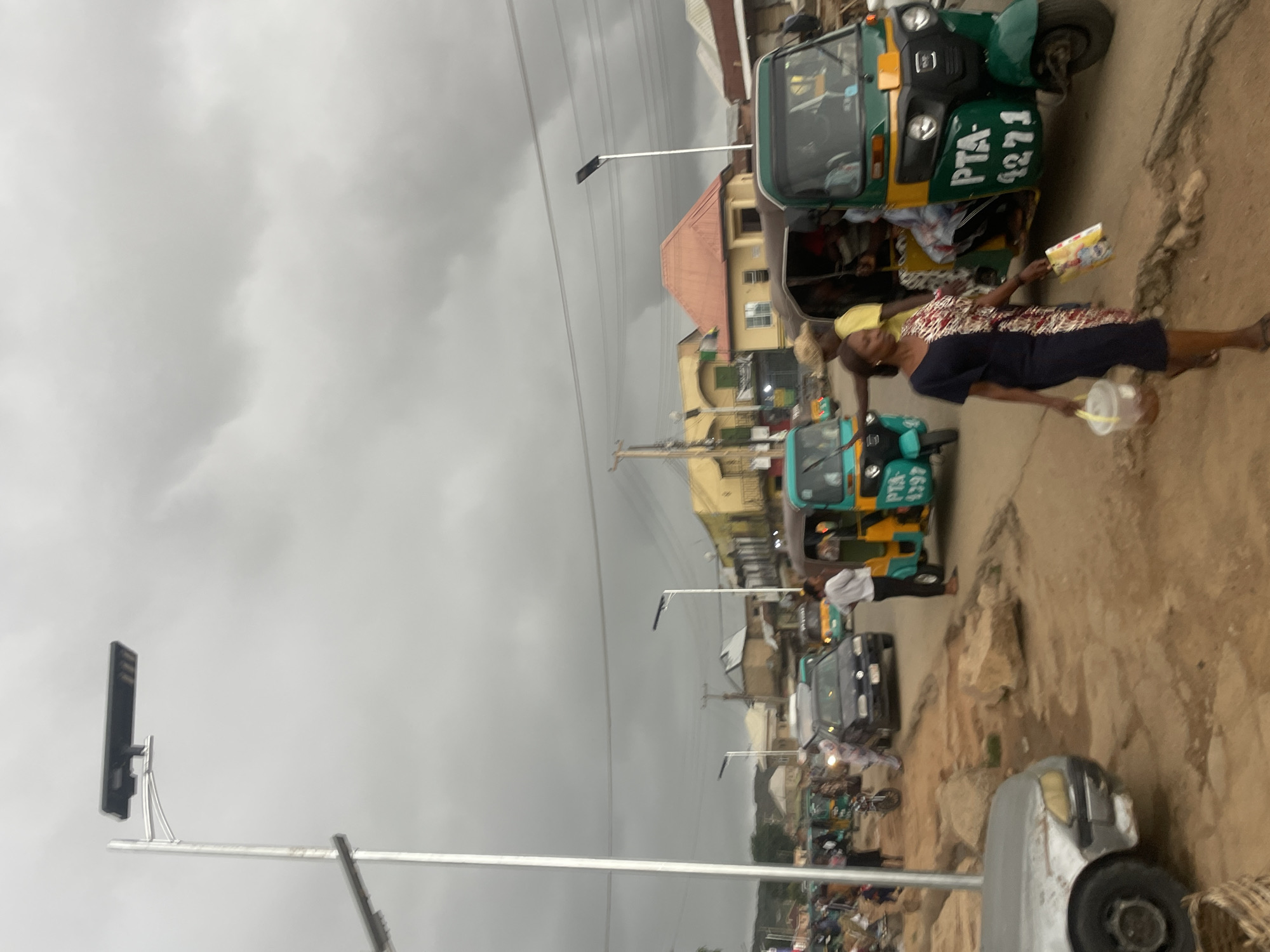 This image depicts a bustling street scene in Jos, Nigeria, under an overcast sky. The road appears unpaved and rocky in parts, lined with various buildings and vehicles suggesting an active urban or semi-urban environment.
In the foreground, a woman wearing a dark blue dress with a patterned upper half is walking. She holds a small, light-colored bucket or container in her left hand and a colorful item, possibly a book or packaged goods, in her right. Another person in a yellow top is partially visible behind her.
Several tricycles, commonly known as Keke Napep in Nigeria, are prominent. A green and yellow tricycle in the upper right section displays "PTA- 1271" on its side. Further down the street, a light blue and yellow tricycle shows "PTA- 4297." These "PTA-" inscriptions likely refer to local transport union or registration details. Other vehicles, including a black car and a partially visible silver-grey car, are also present.
The street is equipped with modern-looking streetlights featuring solar panels, mounted on poles. Overhead power lines are visible against the cloudy sky. The buildings lining the street vary in size and color, suggesting a mix of residential and commercial structures. The activity appears to be typical daily life, with people moving along the street and public transportation operating. The overcast sky indicates daytime with cloudy weather.
