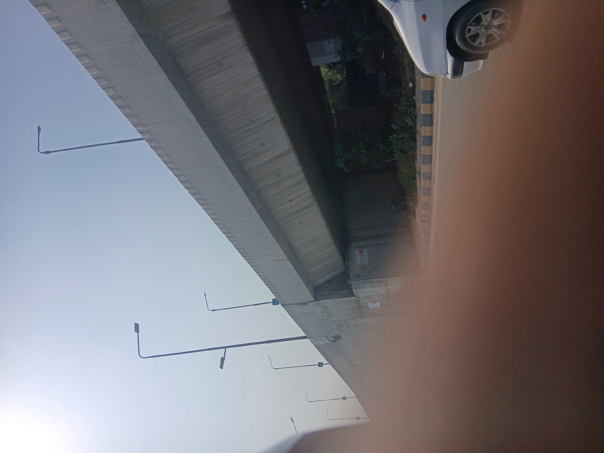 The image shows a concrete overpass and road infrastructure in Peshawar City Tehsil, Pakistan. Several streetlights are visible along the overpass, which appears to be a multi-lane structure. The sky is clear and bright, indicating daytime. Below the overpass, there is some vegetation and greenery. A white car is partially visible in the foreground, driving on the road. The scene is typical of urban infrastructure in Pakistan, with a focus on transportation and public amenities.