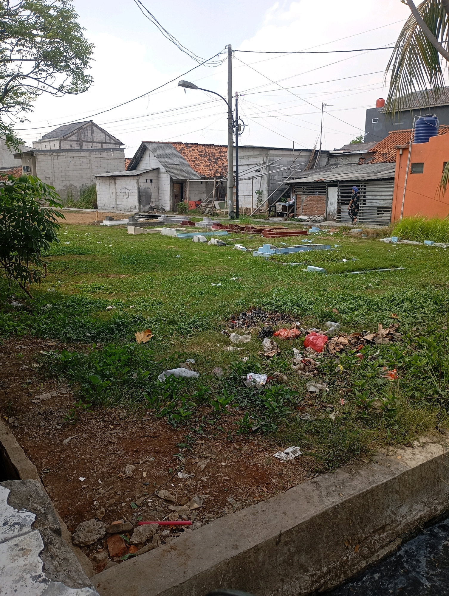 In Babelan, Indonesia, an outdoor scene is depicted. The foreground features a concrete drainage channel with dark, murky water. The bank of the channel is lined with concrete and dirt, littered with small rocks, broken bricks, and debris. Beyond the channel is an area of uneven ground covered in green grass, weeds, and scattered trash, including plastic bags, wrappers, and what appear to be dried leaves.
Further back, a collection of simple, mostly light blue and gray tombstones, some with red trim, are arranged on the grassy expanse, suggesting a cemetery or memorial site. Several structures are visible in the background. To the left, a two-story building with a grey tiled roof and white brick walls stands. Adjacent to it are single-story buildings with corrugated metal roofs and white brick construction, some appearing unfinished. In the center and right of the background, more buildings with red tile roofs and various construction materials are present. A person in dark clothing with a blue head covering is walking past one of these buildings. Overhead, power lines and a street light are visible against a bright sky. A large orange building with a blue water tank on its roof is situated on the far right. The overall environment appears unkempt and somewhat neglected.