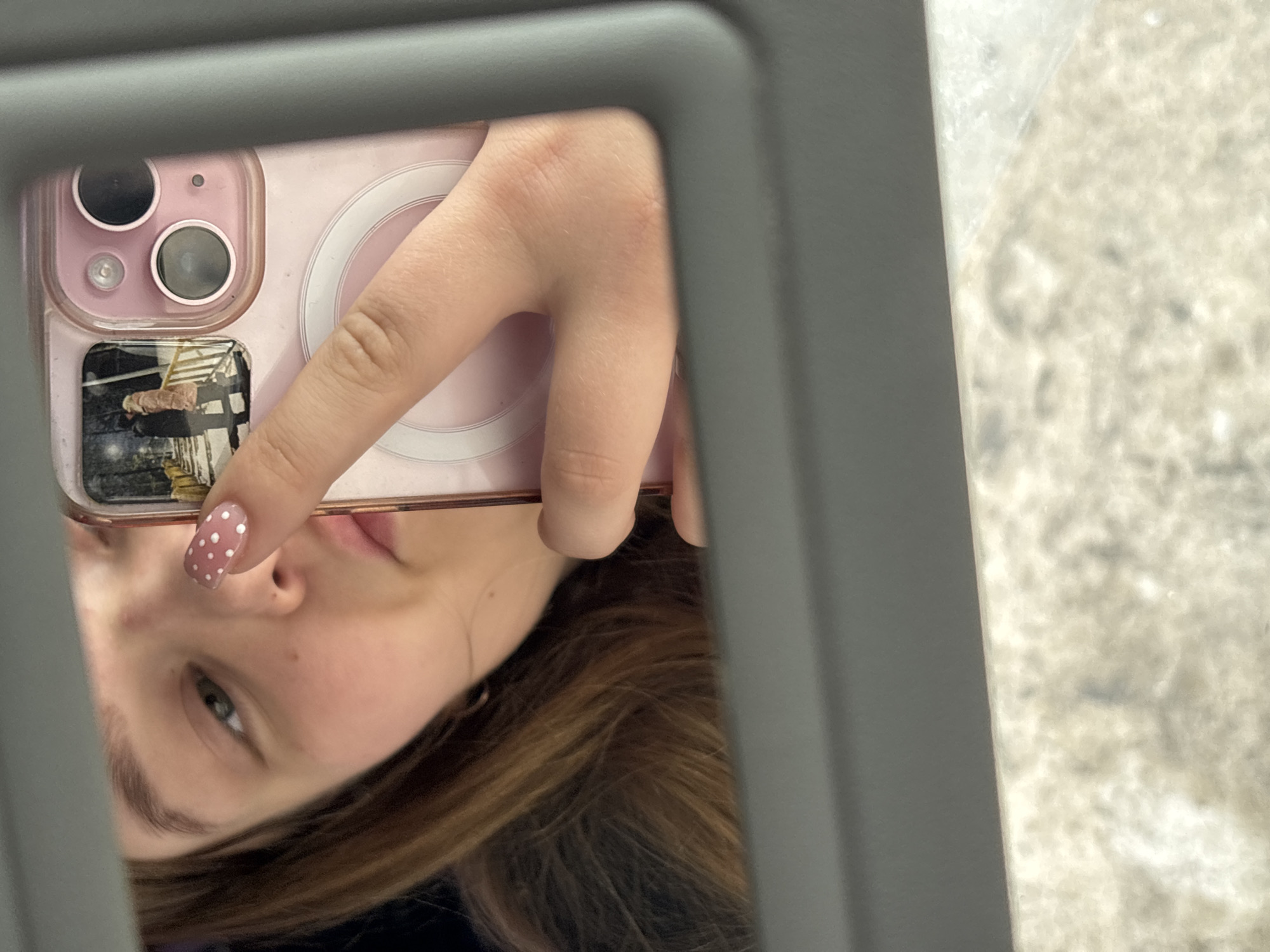 A young person is holding a pink-cased iPhone, reflecting their face in a gray-framed mirror. The background visible outside the mirror is a mottled light-gray surface. The person has light brown hair and fair skin, with one nail painted pink with white polka dots. The phone's case features visible camera lenses and a circular magnetic charging element, and a small photo or sticker on the lower left. The setting is undetermined. There is no information to indicate that this image was taken in Chaikovsky, Russia.