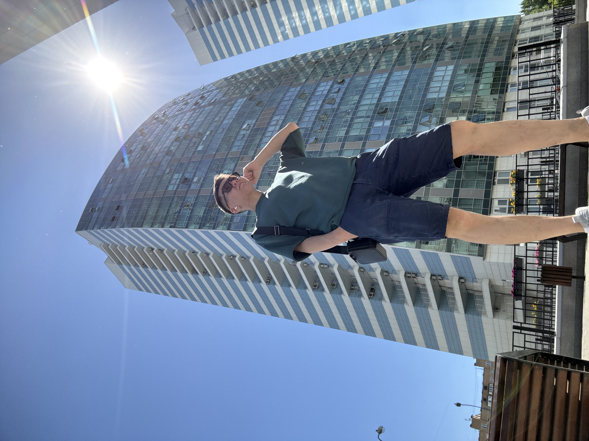 The image depicts a young man posing in a modern urban environment under a bright, clear blue sky. The scene is located in the **LIFE-Varshavskaya Residential Complex area in Moscow, Russia**.
The man, appearing to be of East Asian descent, stands at the center, looking upwards and to his left. He wears dark sunglasses, white wireless earbuds, a dark green short-sleeved t-shirt, dark blue shorts, and light grey sneakers. A black cross-body bag hangs from his left shoulder. His left hand is casually placed in his shorts pocket, while his right hand is raised to his ear, index finger extended towards his temple.
In the background, two prominent modern high-rise buildings dominate the skyline. The larger building to the right is a tall skyscraper with a slightly curved, multi-story glass facade, reflecting the blue sky. Many of its windows are visible, with some partially open. To the left of this building, partially obscured by the man, is another tall residential building characterized by distinctive horizontal bands of light blue/grey and white. This building features multiple balconies with white railings, and numerous external air conditioning units are visible on these balconies. Further in the distant background on the far left, other lower-rise residential structures are faintly visible.
The sun is directly overhead or slightly to the right, causing a strong lens flare and rainbow streaks in the upper right quadrant of the image. The bright sunlight suggests a midday setting. In the foreground, a dark metal fence with planters containing yellow and pink flowers runs along the bottom edge. To the far left, a section of a wooden bench or similar structure is partially visible, along with a black street lamp. The man is standing on a light-colored paved surface. The image is captured from a low angle, emphasizing the height of the buildings and the man's pose against the sky.