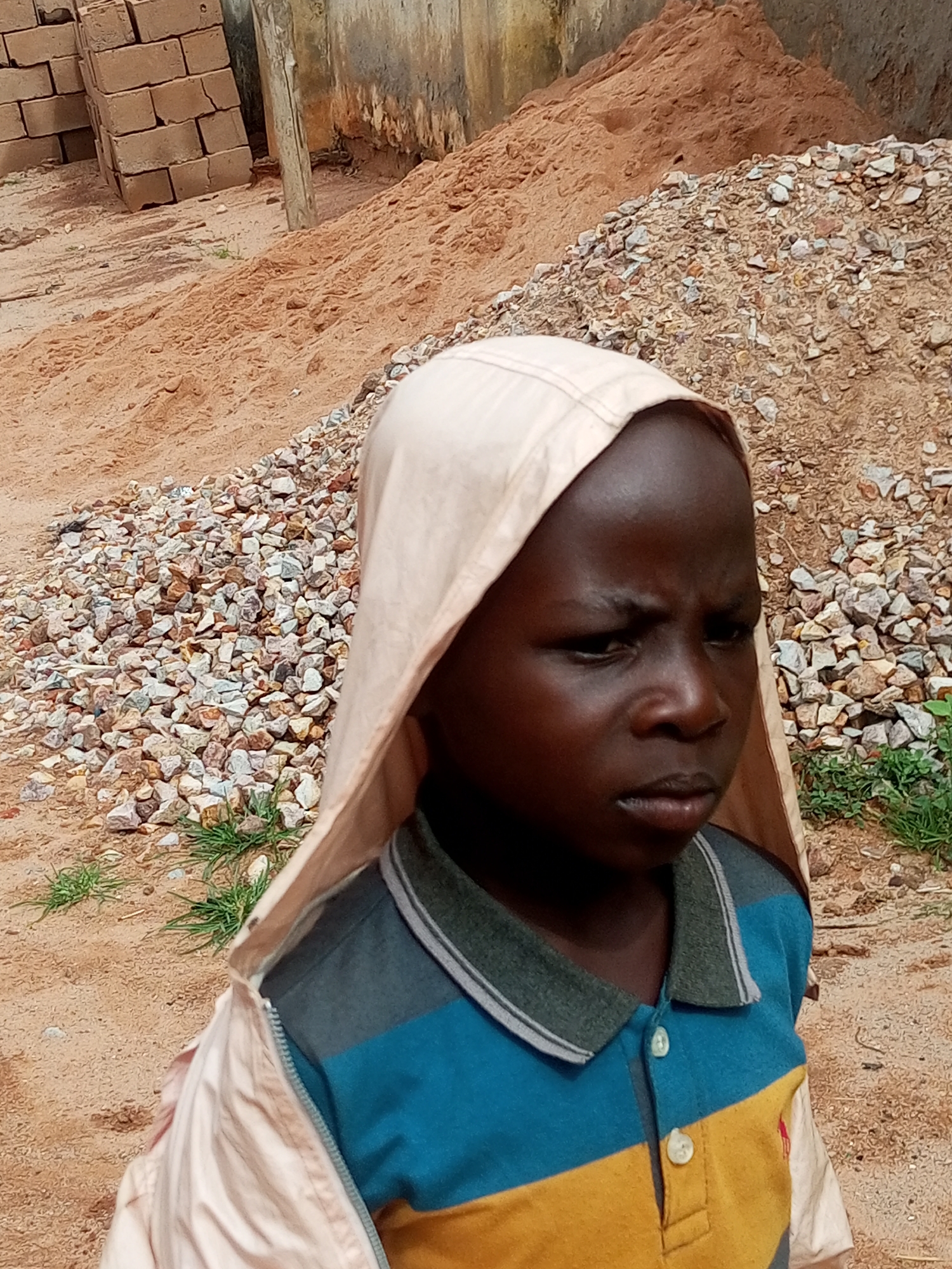 The image shows a close-up of a young boy wearing a light-colored hooded jacket over a collared shirt. The shirt has horizontal stripes of blue, gray, and yellow, with two white buttons at the collar. The boy has dark skin and is looking to his right with a serious expression. In the background, there are piles of sand and gravel, as well as a stack of bricks. The ground is sandy and some sparse green grass is visible. The environment appears to be an outdoor construction or work site. The exact location in Tempure, Nigeria is not specified by visual cues.