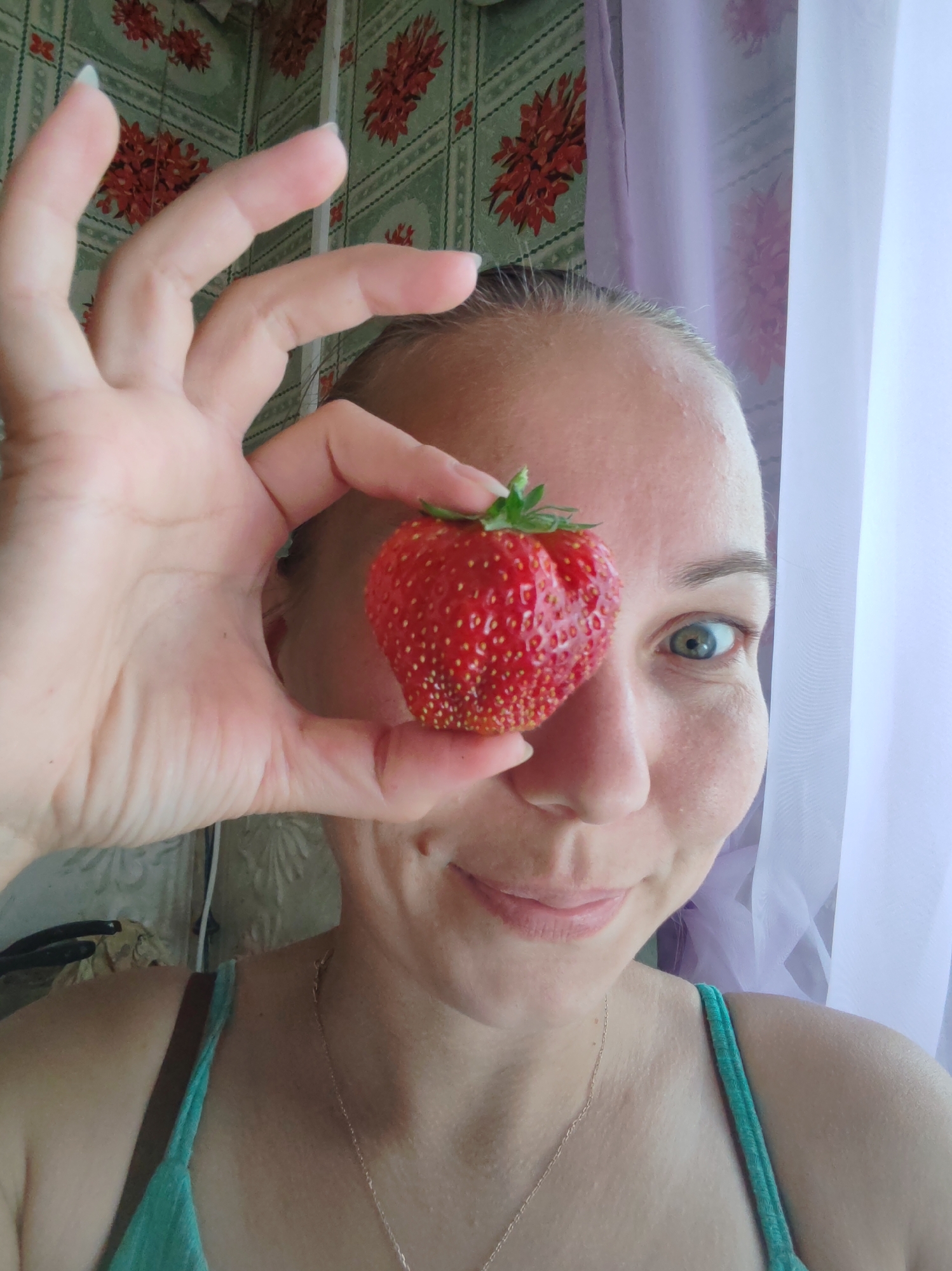 A woman is holding a strawberry in front of her left eye, obscuring it from view. The strawberry is bright red with visible seeds. Her right eye is open and looking towards the camera. She has fair skin with some light freckling on her nose and cheeks. Her mouth is slightly open in a smile. She is wearing a teal-colored tank top. The background appears to be a wall with a floral pattern and sheer curtains. The image quality is slightly blurry. There is no information in the image to suggest a location in the city of Чухлэм, Russia.