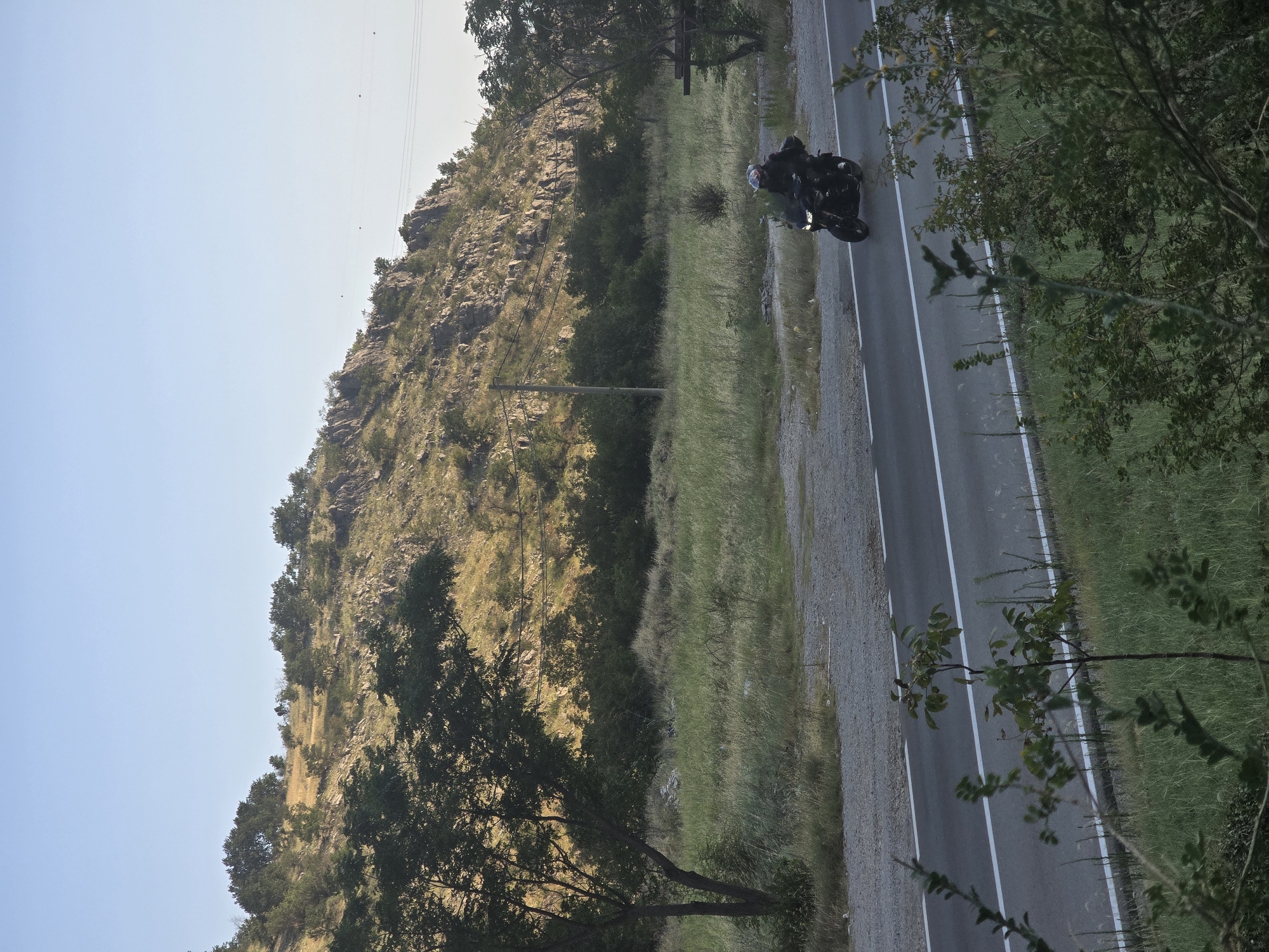 A lone motorcyclist is seen riding on a paved road that cuts through a rural landscape. The road has two white lines marking its lanes. To the left of the road, there is a grassy field with some scattered trees and shrubs. In the background, a rocky, tree-covered hill rises under a clear, pale blue sky. The scene appears to be a highway in a natural, undeveloped area.