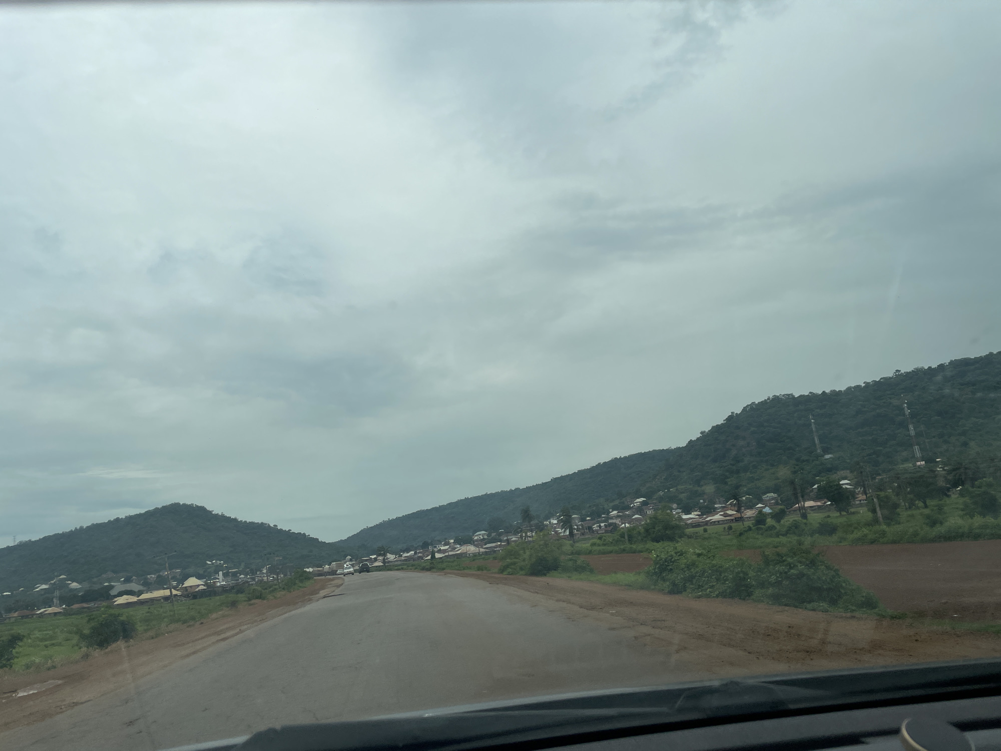 The image depicts a scenic view of a road winding through a landscape with lush green hills and a small village. The sky is overcast with a cloudy, hazy appearance, suggesting it might be early morning or a cloudy day. The road is paved and appears to be a two-lane highway, with a dirt shoulder on the right side.
The village is nestled at the foot of the hills, characterized by numerous buildings with various roof types, including what appear to be corrugated metal and tiled roofs. Some of the structures seem to be residential homes, while others might be public buildings like schools or community centers. There are also several communication towers visible on the slopes of the hills.
The vegetation is dense, with trees and bushes covering the hillsides. In the foreground, there are patches of tilled earth, indicating agricultural activity. A few vehicles are seen on the road, including cars and what looks like a truck, suggesting the road is active with local traffic.
The overall impression is a serene, rural setting in Nigeria, possibly Koton Karifi, given the context. The cloudy weather contributes to a soft, diffused light, creating a calm and tranquil atmosphere. There are no visible people in the image, and no text is present. The scene captures a typical view from a moving vehicle, looking out towards the surrounding countryside.