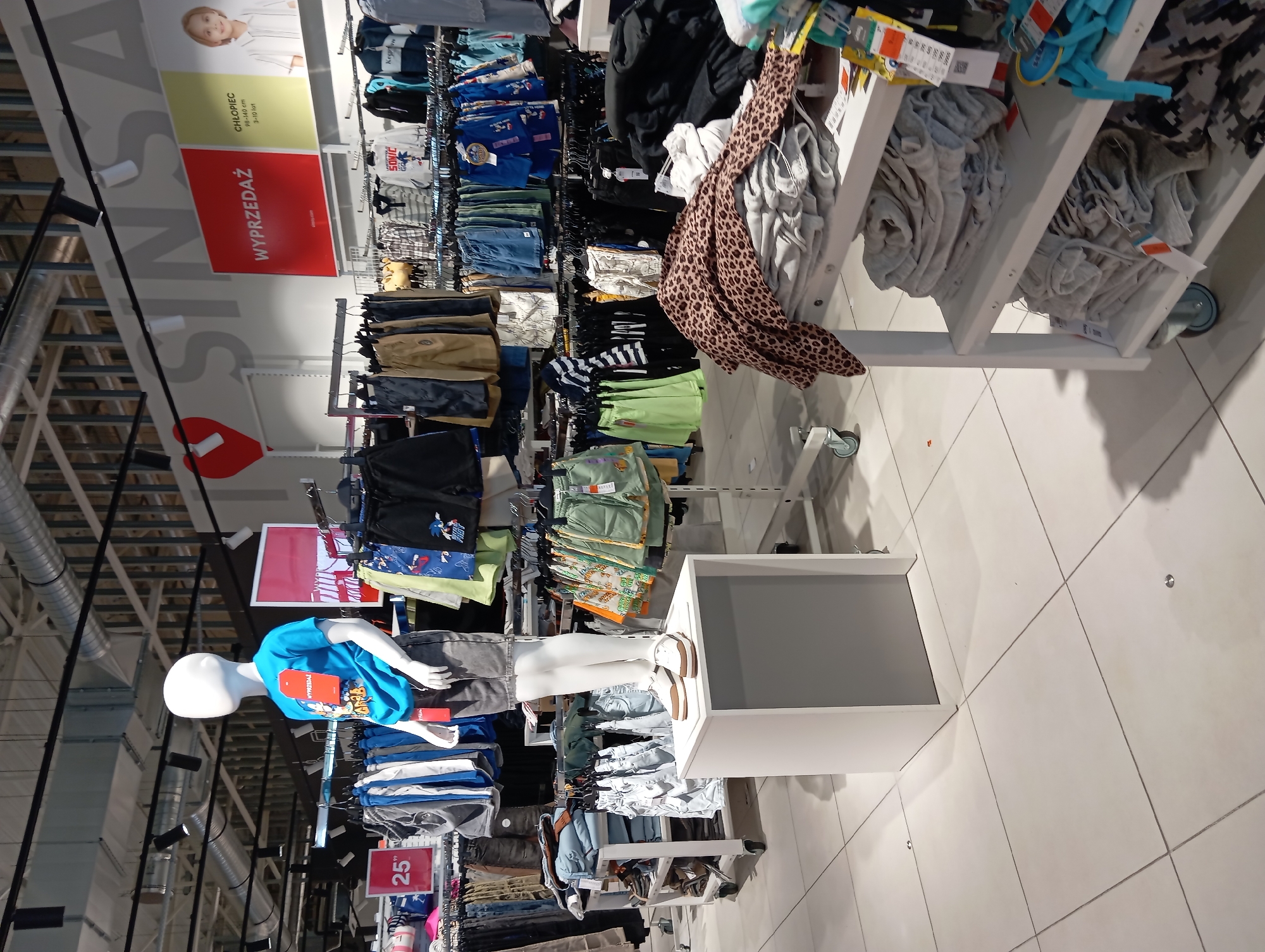The image shows a retail display of children's clothing in what appears to be a store in Wołomin, Poland. The setting is a well-lit retail environment with white tiled floors and industrial-style ceiling elements.
The main subjects are clothing items displayed on racks and shelves, featuring various garments like shorts, t-shirts, and pants. A prominent feature is a child-sized mannequin wearing a blue t-shirt with a graphic design and grey shorts, positioned to showcase the apparel. Nearby, a sign with the text "WYPRZEDAŻ" (Polish for "SALE") in red is visible, indicating a promotional event. Another sign displays "25" followed by a punctuation mark, likely indicating a price or discount.
The overall activity is the presentation of merchandise for sale. There are no discernible people actively shopping in this frame, but the context clearly implies a commercial setting. The lighting suggests daytime, and there are no clues about the weather. The arrangement of clothing and display units is typical of a clothing store, with a focus on children's wear. The brand "SONIC" is visible on some of the clothing items.