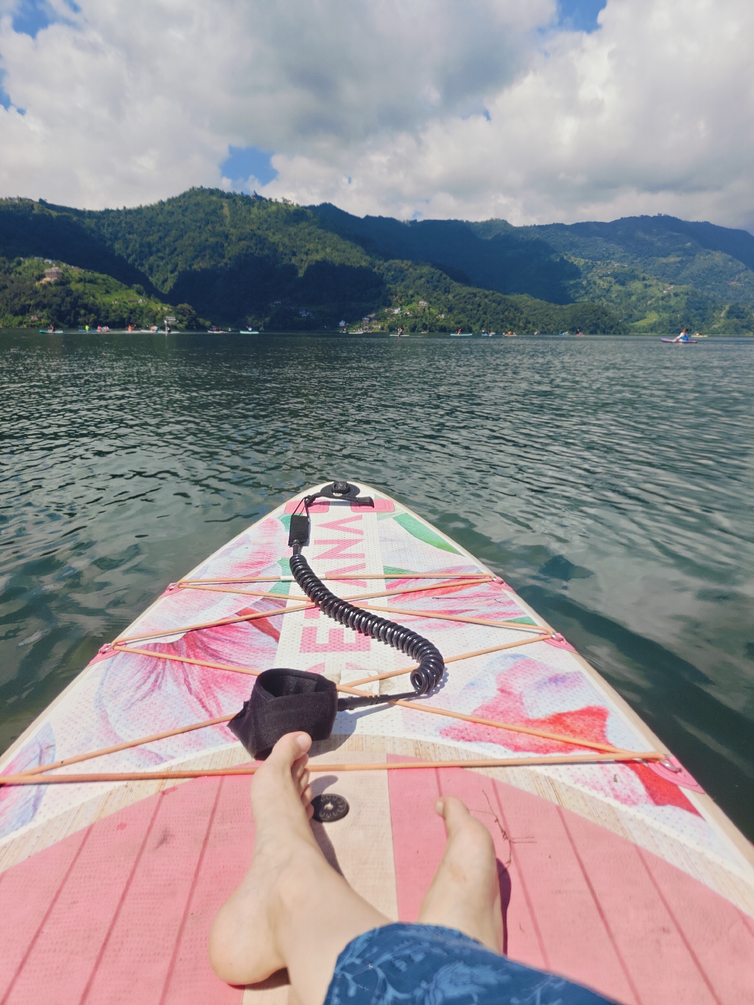 A first-person perspective shot shows a person's feet resting on a stand-up paddleboard on a lake. The paddleboard is decorated with floral patterns and has a coiled leash attached to the person's ankle. In the background, lush green mountains rise from the water, and several other paddleboarders can be seen on the lake. The sky is partly cloudy with patches of blue. The scene is likely on Phewa Lake in Pokhara, Nepal, a popular destination for paddleboarding and enjoying the natural scenery.