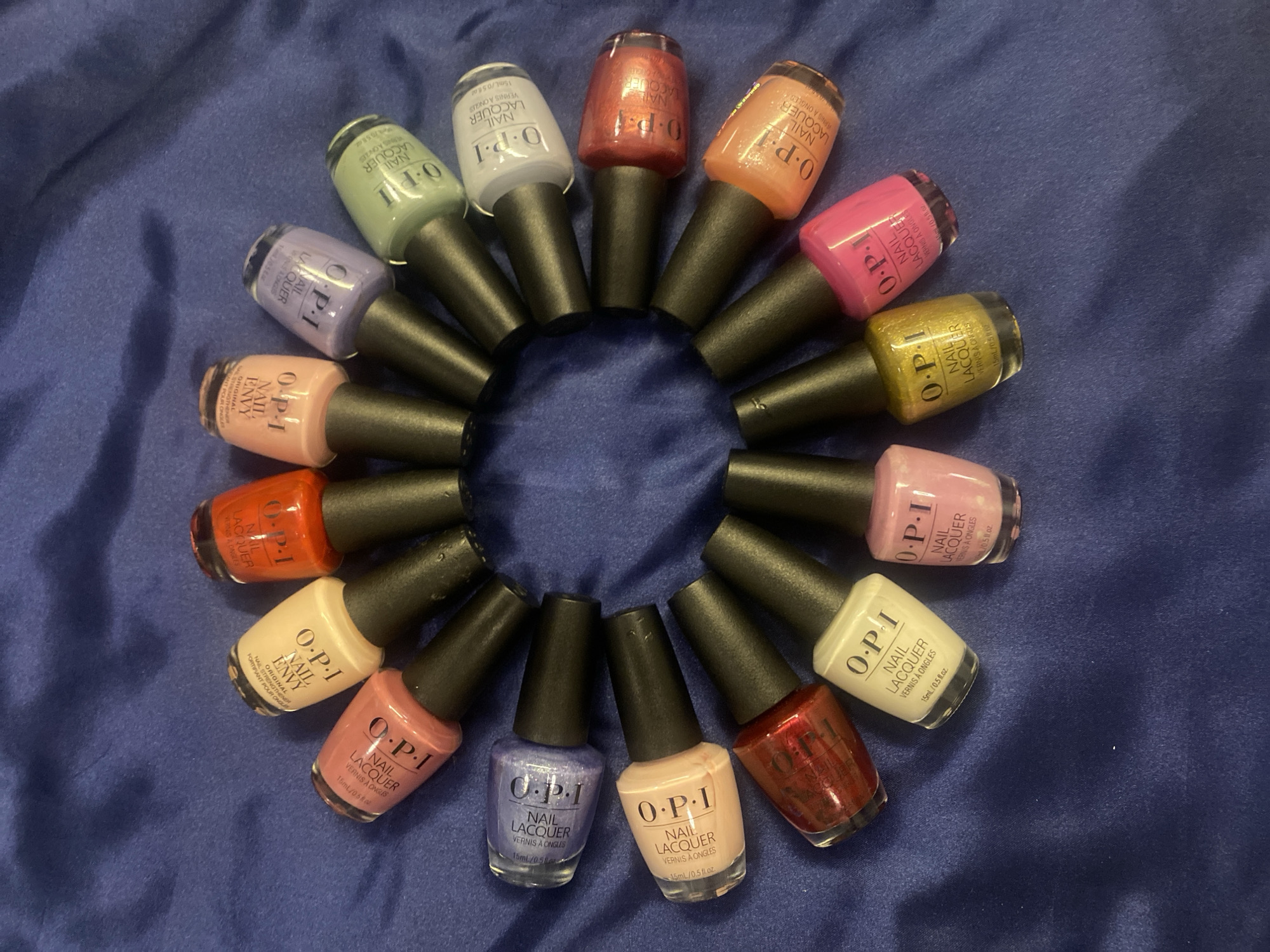 A circular arrangement of 12 OPI nail polish bottles is displayed on a royal blue, textured fabric, resembling satin. The bottles are oriented with their caps pointing inwards towards the center of the circle. The fabric has visible folds and creases, creating shadows and highlights.
The nail polish colors, starting from the top and moving clockwise, appear to be: a peachy nude, a vibrant orange-red, a pale peach, a light lavender, a mint green, a pale sky blue, a metallic coral, a bright orange, a hot pink, a metallic gold, a light pink, and a deep red with subtle shimmer. Each bottle features the "O.P.I NAIL LACQUER" label in black text on a white background, with the volume "15ml / 0.5 fl.oz." and "VERNIS A ONGLES" also printed.
There are no people present in the image. The scene is indoors, on a flat surface. The lighting suggests a direct light source from above, casting shadows from the bottles onto the fabric. The arrangement forms a cohesive collection, possibly a display or personal collection of OPI nail polishes. The context of Kuala Lumpur, Malaysia, is not visually indicated in the image itself.
