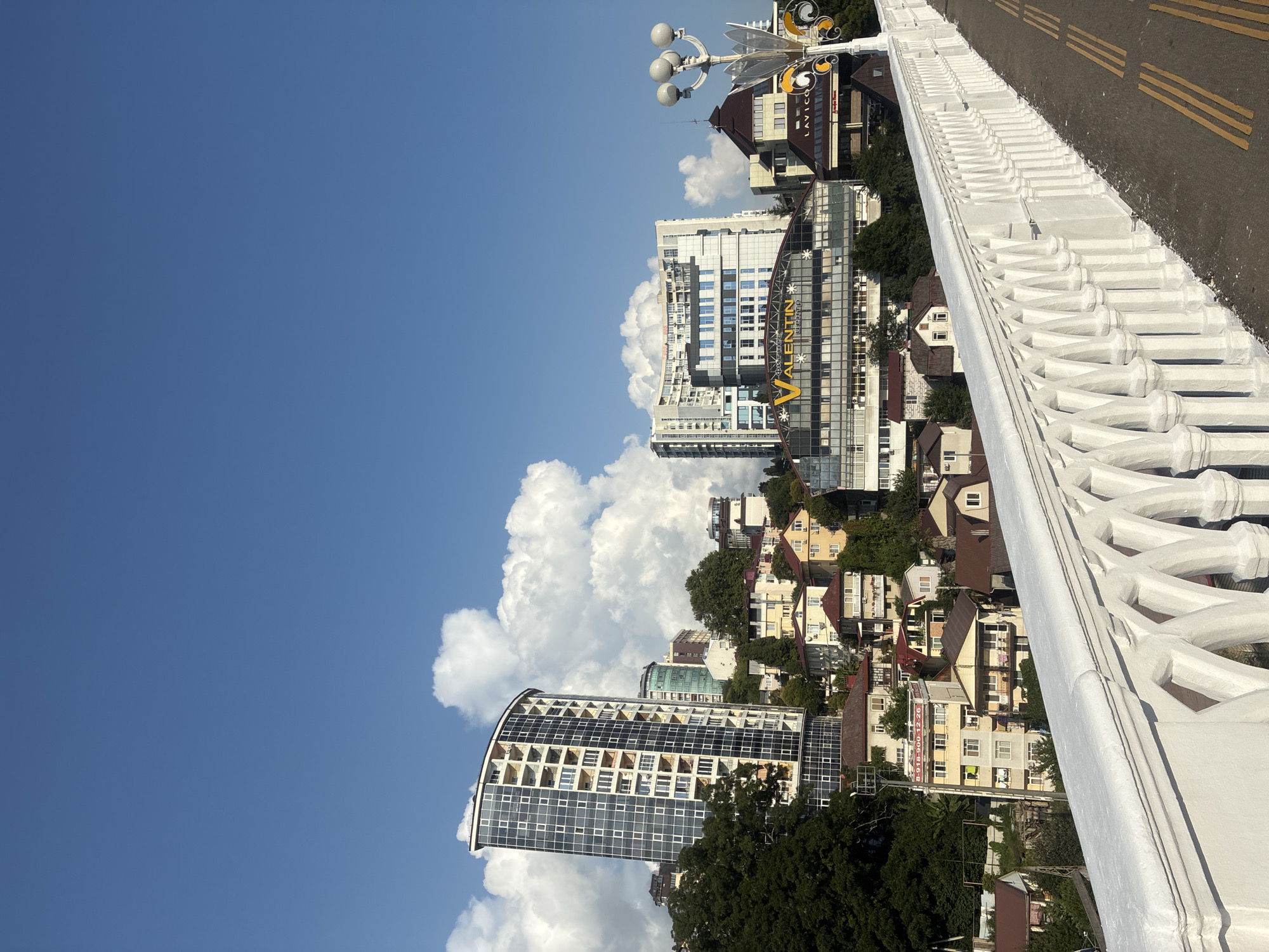 The image shows a view of buildings in Sochi, Russia, with a bright blue sky and large white clouds. In the foreground, there is a white ornate railing along a path or bridge. Several modern buildings are visible, including a tall, curved skyscraper with many windows, and a longer, multi-story building with a curved glass facade and a prominent yellow "VALENTIN" sign. Smaller residential buildings with red and yellow roofs are nestled on a hillside behind the modern structures. Trees are interspersed among the buildings. A lamppost with multiple globes stands on the right side of the image, near the edge of the path. The overall scene depicts an urban landscape with a backdrop of natural elements.