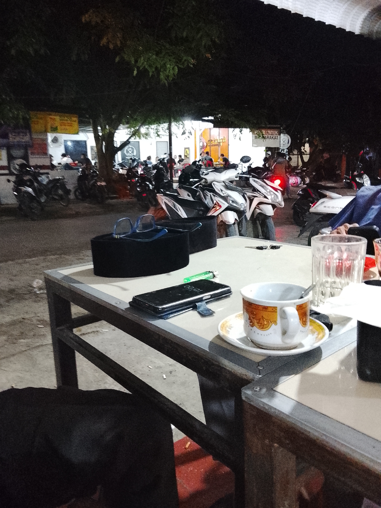 The image shows a table at an outdoor café in Lhokseumawe, Indonesia at night. The table holds a cup of coffee, a saucer, eyeglasses, a black peci hat, a smartphone, a lighter, a glass, and what appears to be an ashtray or a coaster. Behind the table, there are several motorcycles parked in front of the café. People are visible in the background, both inside and outside the building. Overhead illumination is provided by light fixtures under the roof of the building and streetlights. There are trees in the middle ground. The setting is a casual, public space.