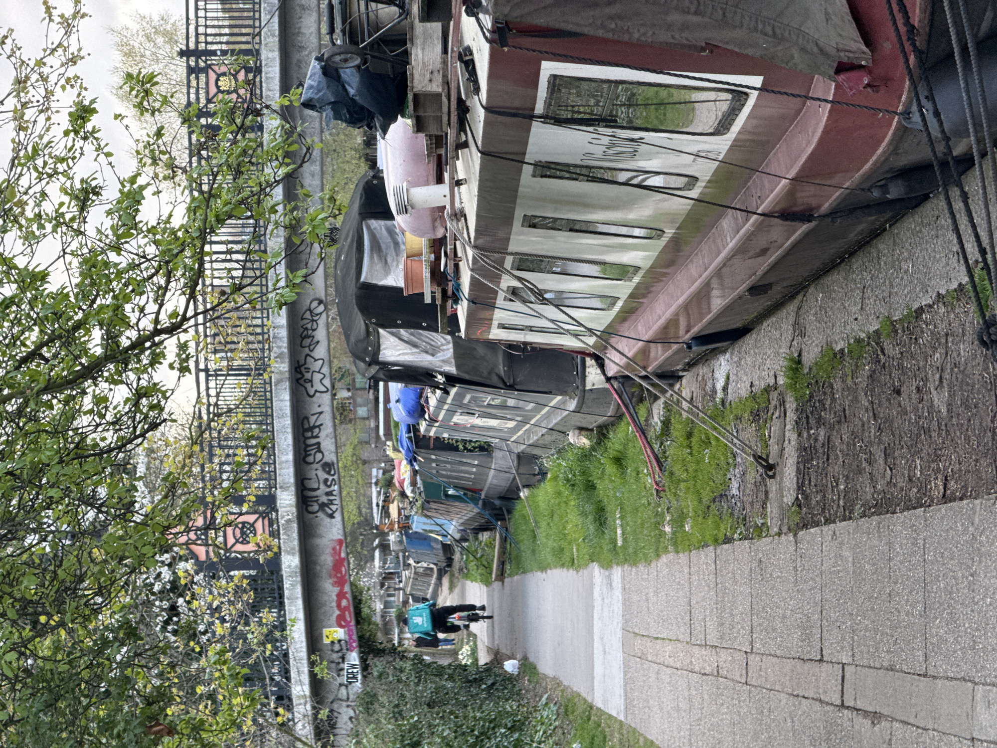 The image depicts a row of narrowboats moored along the Regent's Canal in London, United Kingdom. A towpath runs alongside the canal, with a delivery cyclist, identifiable by the "Deliveroo" bag, riding along it. To the left, a concrete post covered in graffiti rises into the background, with trees and foliage adding greenery to the scene. The narrowboats are various colors and show signs of habitation.
