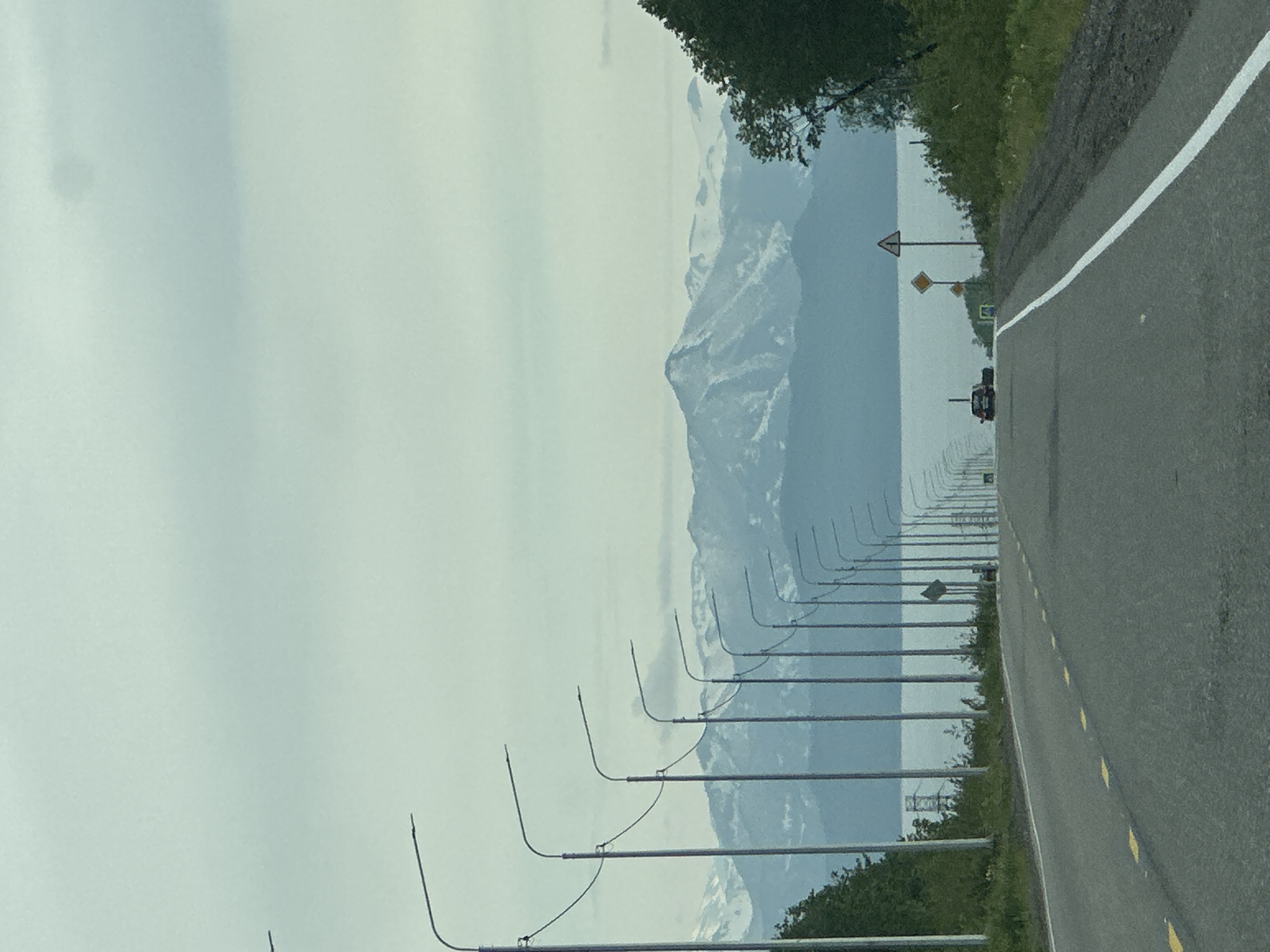 This is a photograph taken from a vehicle traveling on a paved road that appears to be a highway or main thoroughfare in Petropavlovsk-Kamchatsky, Russia. The road is in good condition with clear lane markings and yellow reflective markers. The sky is overcast with a muted, cool tone.
On the left side of the road, a long line of streetlights with curved arms is visible, stretching into the distance. Beyond the streetlights, a body of water, likely the ocean or a large bay, is present. In the background, a range of snow-capped mountains rises dramatically, their peaks obscured by the cloudy sky. The mountains have a rugged, textured appearance.
On the right side of the road, a dense growth of green trees and vegetation lines the shoulder. Further into the distance, more streetlights are visible on the right side of the road, following the curve of the asphalt.
There are a few vehicles present: a dark-colored car is visible in the distance on the right lane of the road, and another vehicle, possibly a pickup truck, is also in the distance on the left lane. Two road signs are visible on the right side of the road; one is a triangular warning sign with a red border, and below it is a diamond-shaped yellow sign with a symbol. The scene suggests an approach to or departure from a populated area, given the presence of streetlights and the quality of the road, set against a backdrop of natural, mountainous terrain.