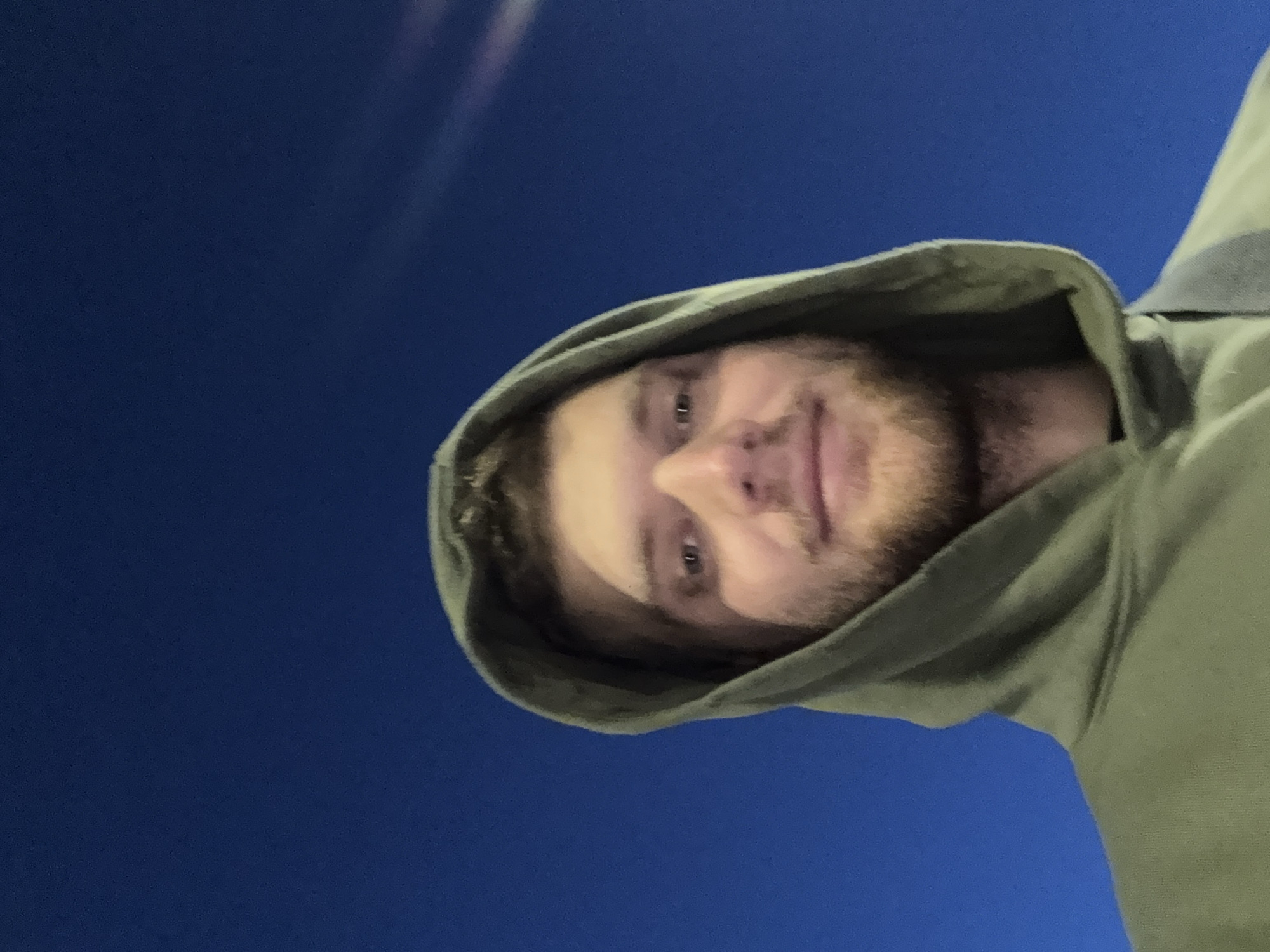 A man with fair skin, a beard, and a hooded sweatshirt is looking directly at the camera. The background appears to be a dark blue, possibly a night sky. The photo is taken from a low angle, looking upwards at the man. The photo does not contain enough information to place this image in Springfield, United States of America.