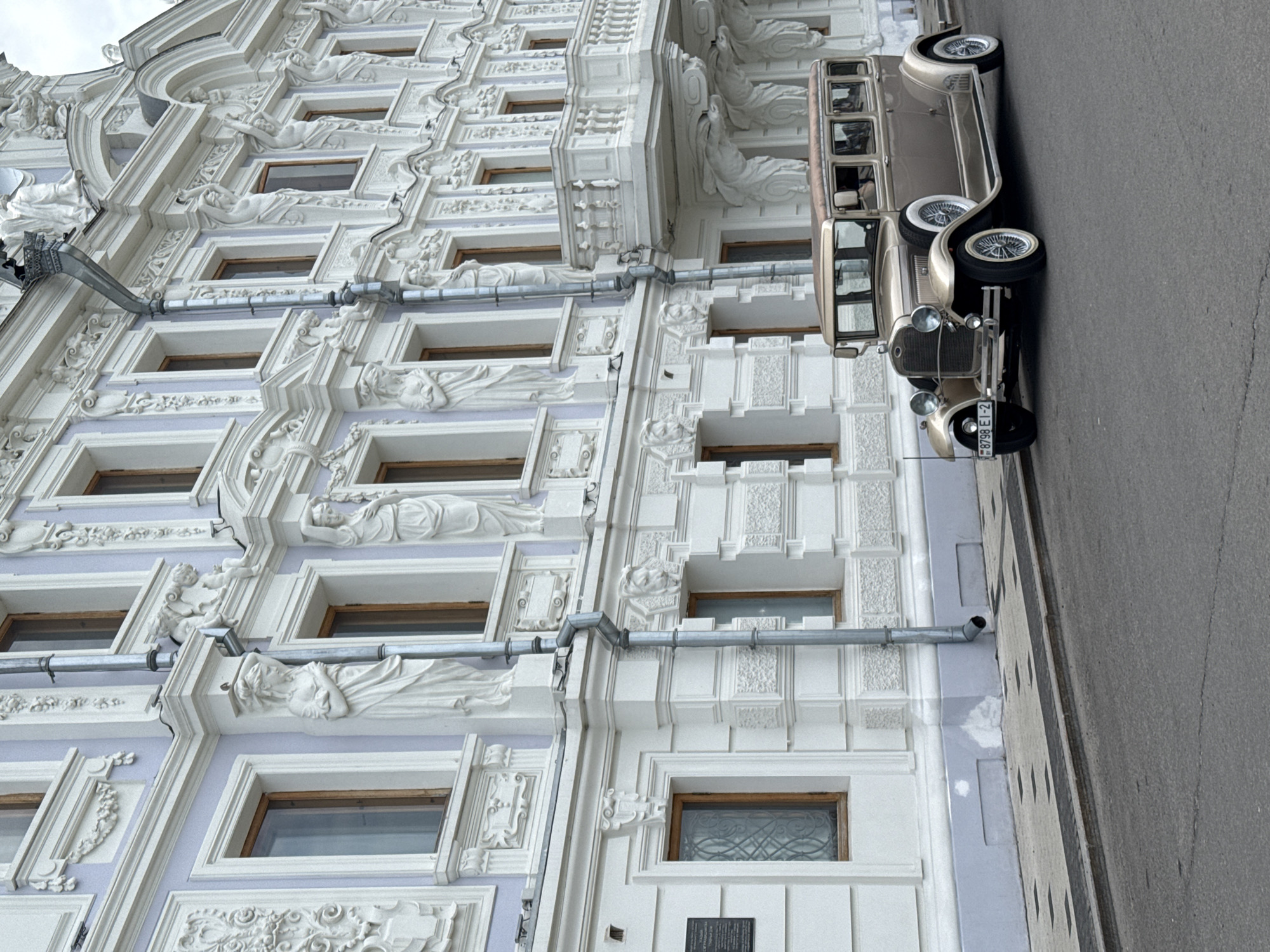 The image depicts a vintage tan-colored car parked in front of a highly ornamented building in Nizhny Novgorod, Russia. The building features elaborate white architectural details, including statues and intricate window frames, against a pale blue background. A metallic drainpipe runs vertically down the side of the building. The car is positioned parallel to the building's facade, with its front end facing toward the top of the image. The pavement is visible on the right side of the frame.