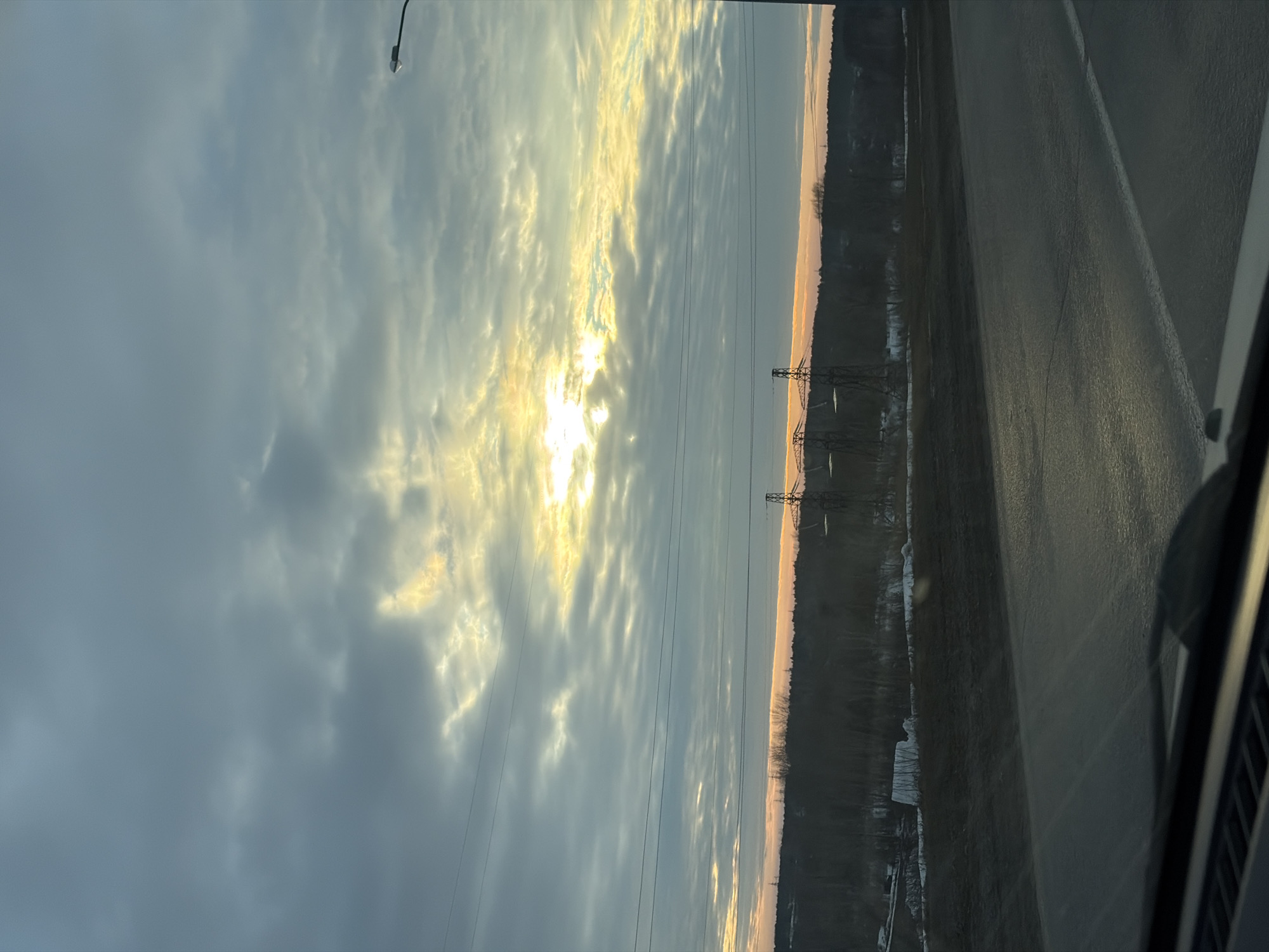The scene depicts an outdoor landscape observed from the interior of a vehicle, likely in the city of Chaikovsky, Russia. The sky, occupying the upper two-thirds of the image, is heavily overcast with stratocumulus clouds, predominantly dark grey and light grey. A bright, yellow-orange sunlight penetrates a central band of clouds, indicating either sunrise or sunset. Horizontal power lines stretch across the mid-upper portion of the sky.
Below the sky, a dark, treeline-dominated horizon is visible, accented by a subtle orange-pink glow. The landscape beneath the treeline consists of dark, possibly muddy or dormant ground, with scattered patches of white snow suggesting a cold season. Two prominent metal lattice power line towers are visible against the horizon, one on the right and another partially visible further left.
In the immediate foreground, the perspective is from inside a vehicle, revealing the dark dashboard, a windshield wiper blade on the right, and what appears to be a vent on the left. A dark asphalt road runs across the lower third of the image, showing a white solid line on its right edge. A black streetlight pole with a fixture extends into the upper right corner of the frame. No individuals are present in the image.