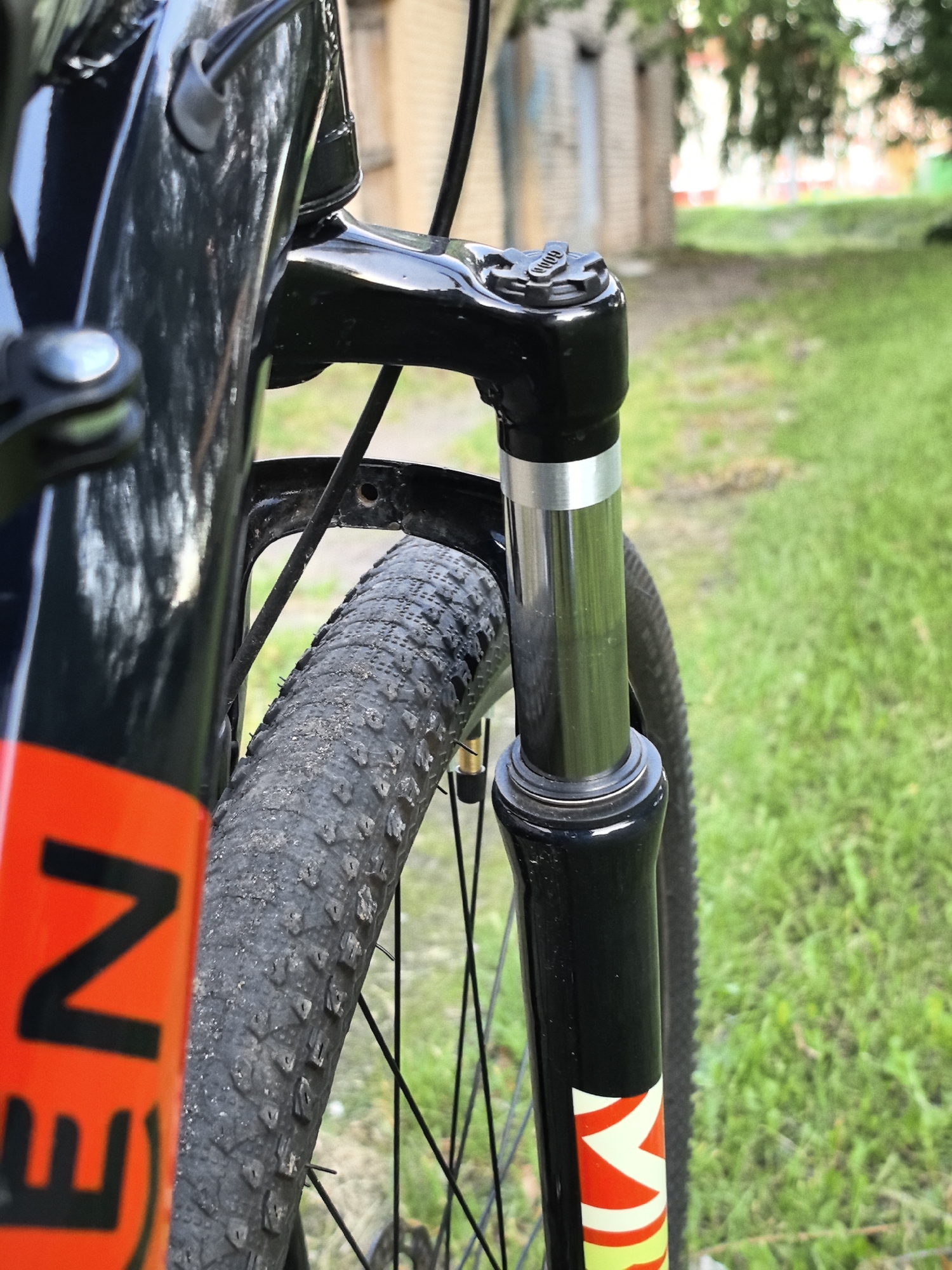 A close-up, eye-level, outdoor shot focuses on the front suspension fork and wheel of a black bicycle. The bicycle is positioned on a grassy area, with a blurred background of trees and buildings, suggesting an urban park or a residential area. The bicycle's frame has a prominent orange stripe with black lettering that reads "EN". The tire is black with visible tread, and it appears to be dusty. The suspension fork is black with a silver band and a black cap on top. The lighting is natural daylight, with soft shadows. No people or animals are present in the image.