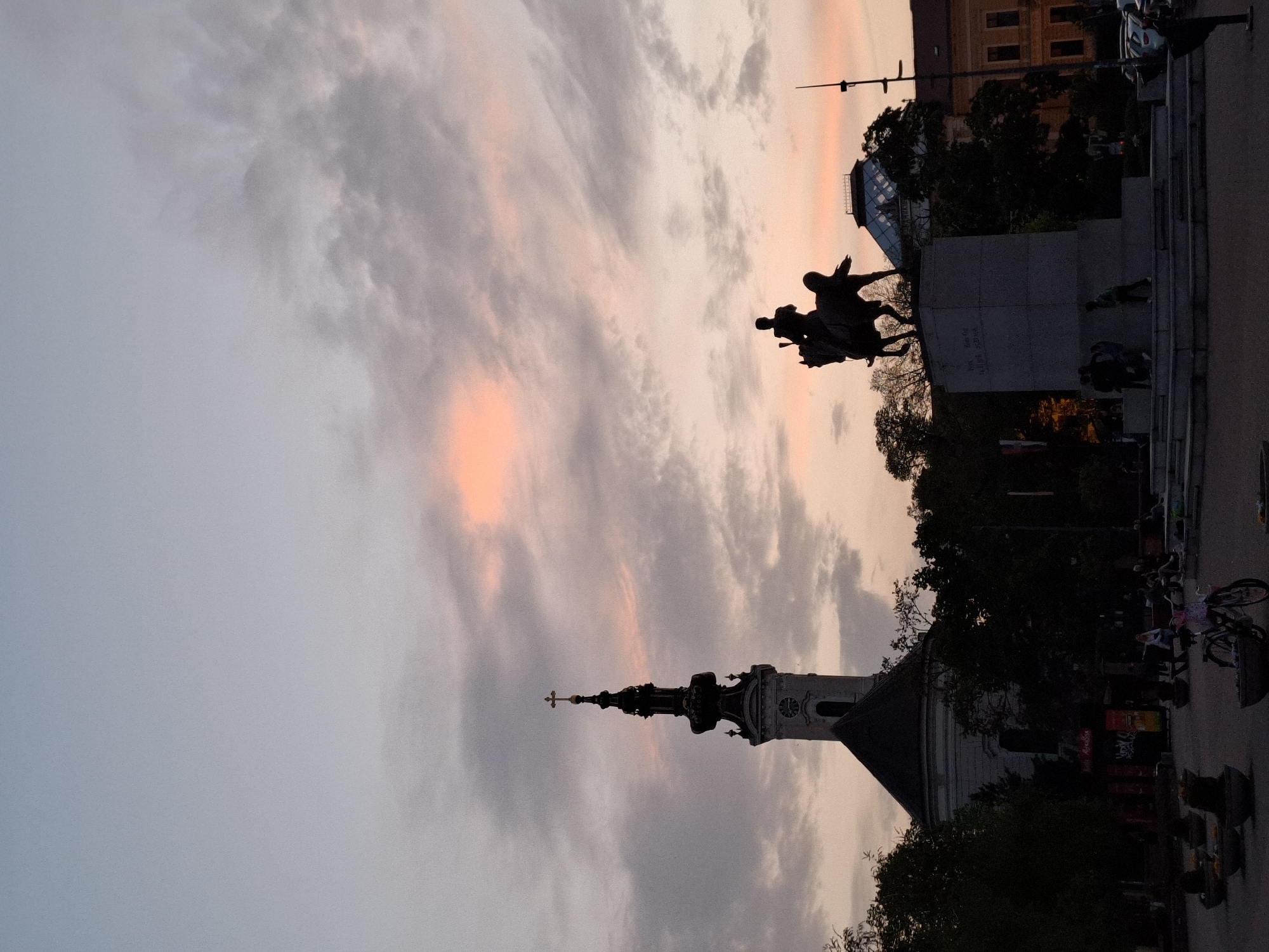 At dusk in Novi Sad, Serbia, a partially silhouetted Orthodox church steeple rises to the left of a statue of a man on horseback. To the right of the statue, a building is seen as well as cars parked along the side of the street. On the plaza near the statue, people appear to be standing and sitting.