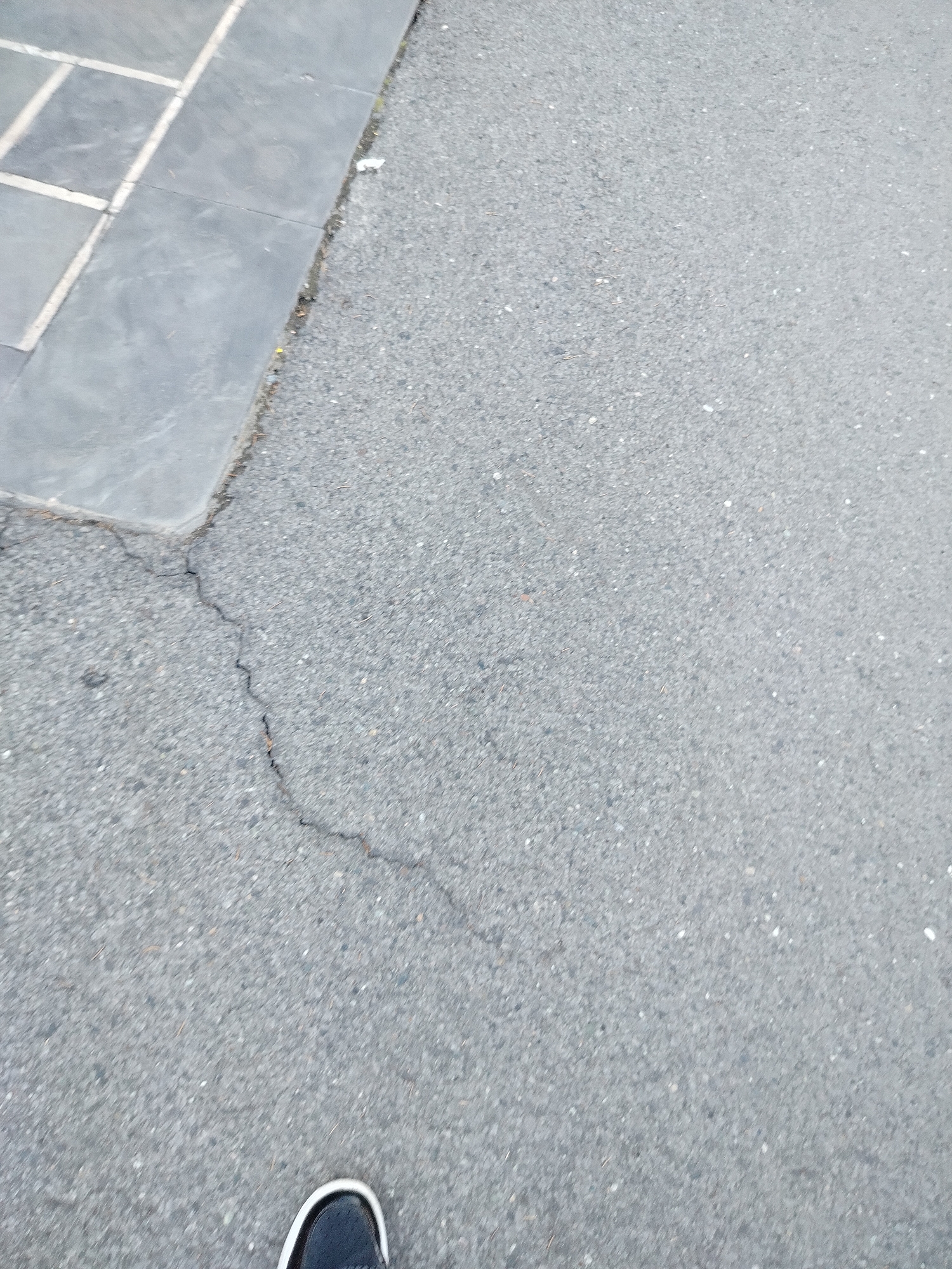 The image shows a section of asphalt pavement with a crack running through it. A portion of a sidewalk made of grey stone tiles is visible in the upper left corner. The toe of a black and white shoe is visible in the bottom of the frame. The location is outdoors, likely a street in Yerevan, Armenia, but there are no specific elements confirming this location.