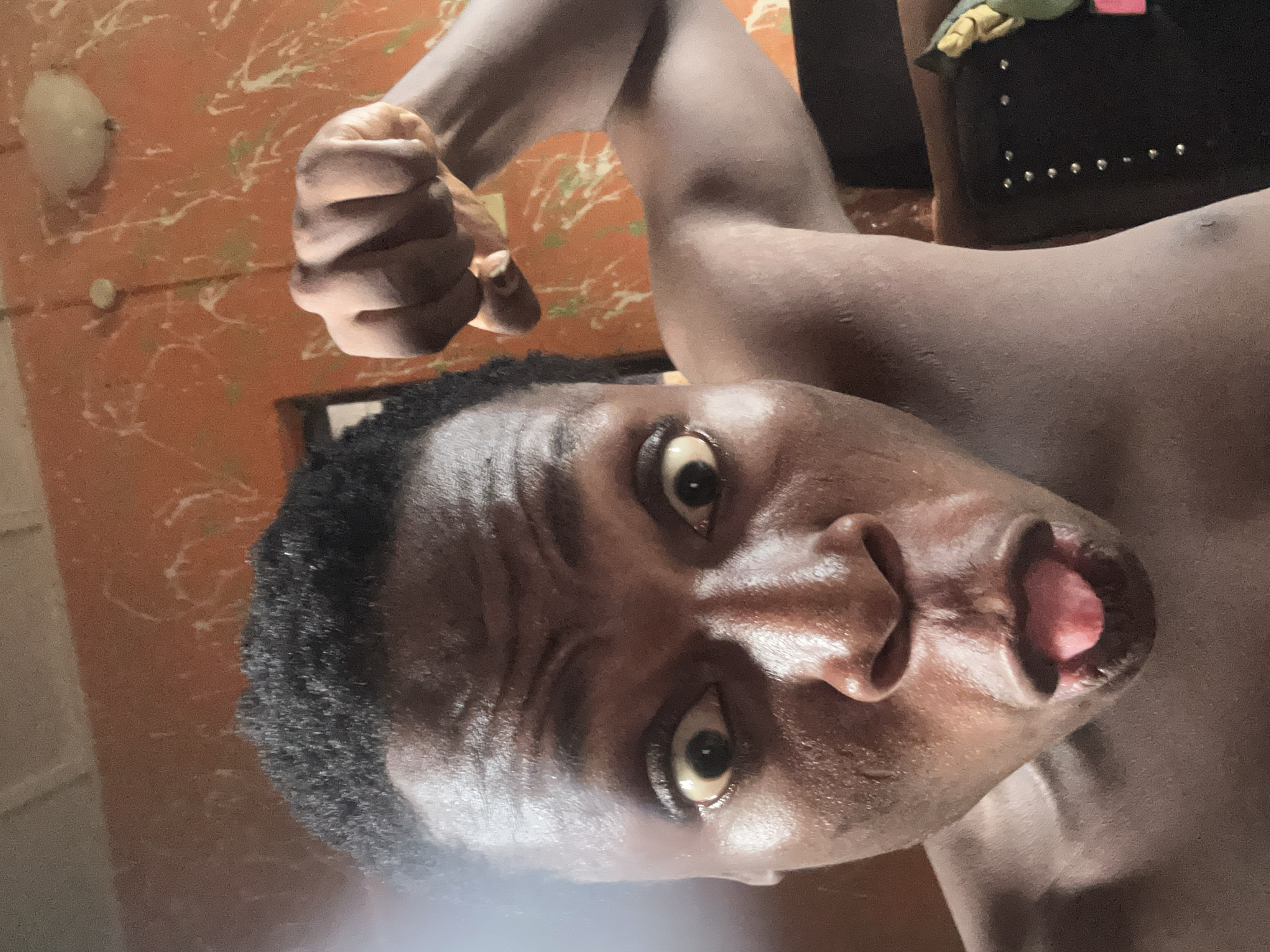 A close-up, tilted image shows a dark-skinned person with short, dark hair, posing with a raised fist. The person's eyes are wide, and their mouth is slightly open. The background is an orange wall with white splatters and a light fixture. To the right, part of a black item with silver studs is visible. The location is an indoor setting.