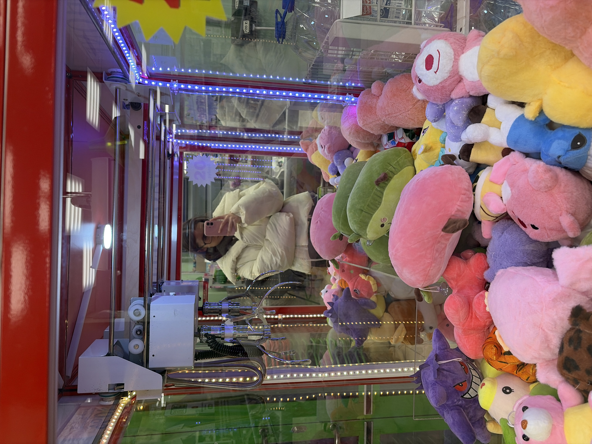 A vertically oriented photograph captures the interior of a claw machine filled with a dense collection of colorful plush toys. The machine features bright red framing and is illuminated by strips of blue LED lights and white lights along its edges.
Inside, a multitude of stuffed animals and objects are piled high. Dominant colors include various shades of pink, green (some resembling avocados), yellow, blue (one character resembling Stitch), and purple (one resembling Gengar). Other brown, orange, and light-colored plushies are also visible, creating a vibrant assortment of prizes.
Reflected clearly in the glass front of the machine is a person, likely the photographer, holding a smartphone to take the picture. The person is wearing a light-colored puffer jacket. Their reflection appears sideways relative to the plush toys, suggesting the camera was held horizontally while capturing the vertically oriented machine. The metal claw mechanism, with its three prongs, is visible on the left side of the machine, positioned above a green base.
The setting is an indoor entertainment venue, likely an arcade, located in Chaikovsky, Russia. Visible text includes "220 В" (220 V) on a sticker inside the machine, indicating electrical voltage. The number "20" is also seen on a yellow star-shaped sticker, potentially indicating a price or prize number. Faint and blurry Cyrillic text is discernable in the reflections on the upper glass surfaces. The image conveys a sense of playful engagement with an arcade game.