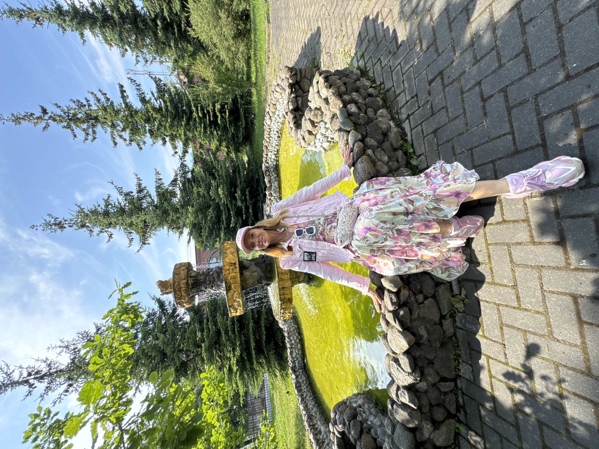 A woman with blonde hair, wearing a pink baseball cap, pink jacket, and a long floral dress, sits on the edge of a fountain. The fountain's water is a bright, almost neon yellow-green, and the fountain itself is made of stacked stone tiers with a decorative element at the top. The fountain is surrounded by a low stone wall. The woman has a light-colored purse with a floral pattern slung across her body. She is wearing pink sneakers. The fountain is situated in an outdoor setting with a paved walkway made of rectangular bricks in front of it. In the background, there are mature evergreen trees and some leafy green trees. The sky is blue with wispy white clouds. The location is identified as a park in Lomonosov, Russia, near the Lomonosov Sea Hydrographic Service Station.