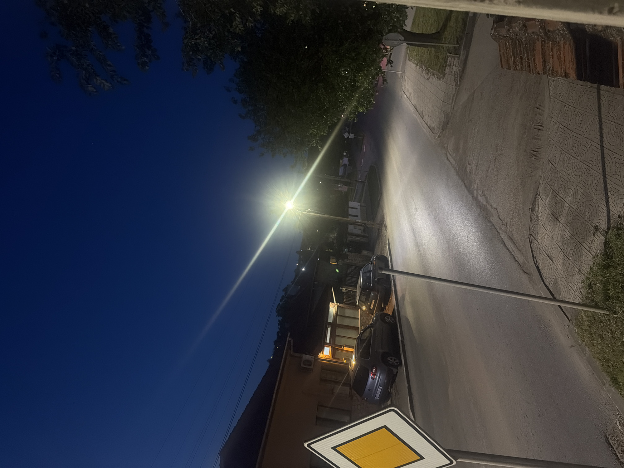 The image displays a night scene of a street in what appears to be a town or village, likely Iskar, Bulgaria, as suggested by the location context. The sky is a deep, dark blue, indicating either early evening or late night. Lush green foliage from trees is visible along the top and upper-left edge of the frame.
A powerful street light dominates the left side of the composition, casting a bright, elongated beam of light across the asphalt road surface. This illumination creates a strong contrast with the dimmer areas of the scene. Along the left side of the road, two dark-colored cars are parked. The rear of the car closer to the foreground shows a white license plate, but the characters are too indistinct to read precisely. Behind the parked cars, a building with several illuminated windows suggests a residential or commercial structure.
On the right side of the road, a patterned sidewalk or pavement runs parallel to the street. In the lower right foreground, a clear yellow diamond-shaped road sign with a white border is prominently displayed on a metal pole, indicating a priority road. This sign is characteristic of European road signage. Faint figures of people can be discerned in the distance, near the base of the street light, suggesting some pedestrian activity. The overall atmosphere is quiet, and the weather appears clear. No specific event or major activity is unfolding.
