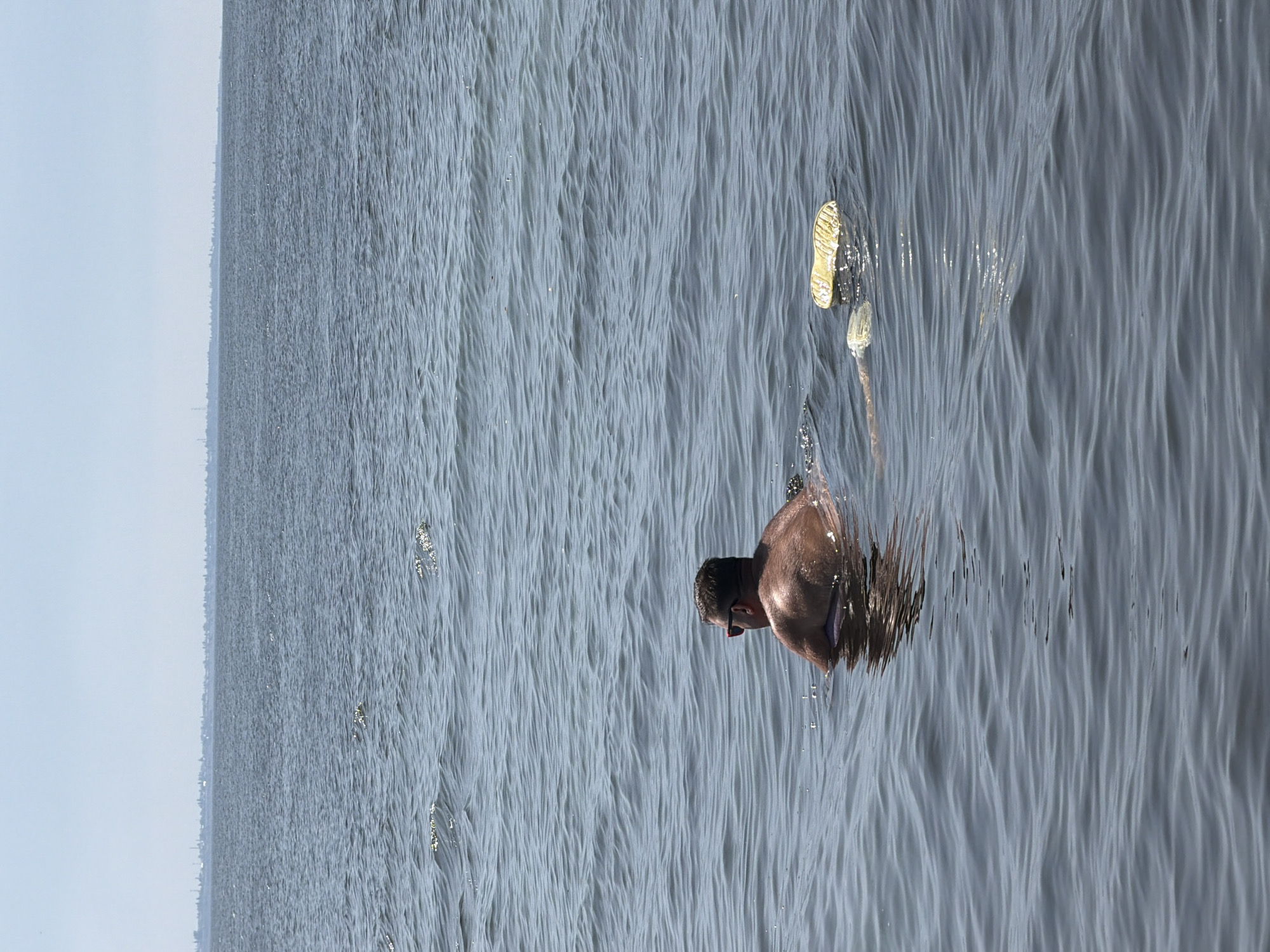 A man wearing sunglasses is swimming in the water. He is in the middle of the frame, facing away from the camera and towards a distant shoreline. A yellow object, possibly a flotation device or a shoe, is floating in the water to his right. The water is calm with small ripples, and the sky is clear. The distant shoreline shows a hazy outline of buildings. The image appears to have been taken from a boat or another watercraft. The location is the Volga River near Kamyshin, Russia.