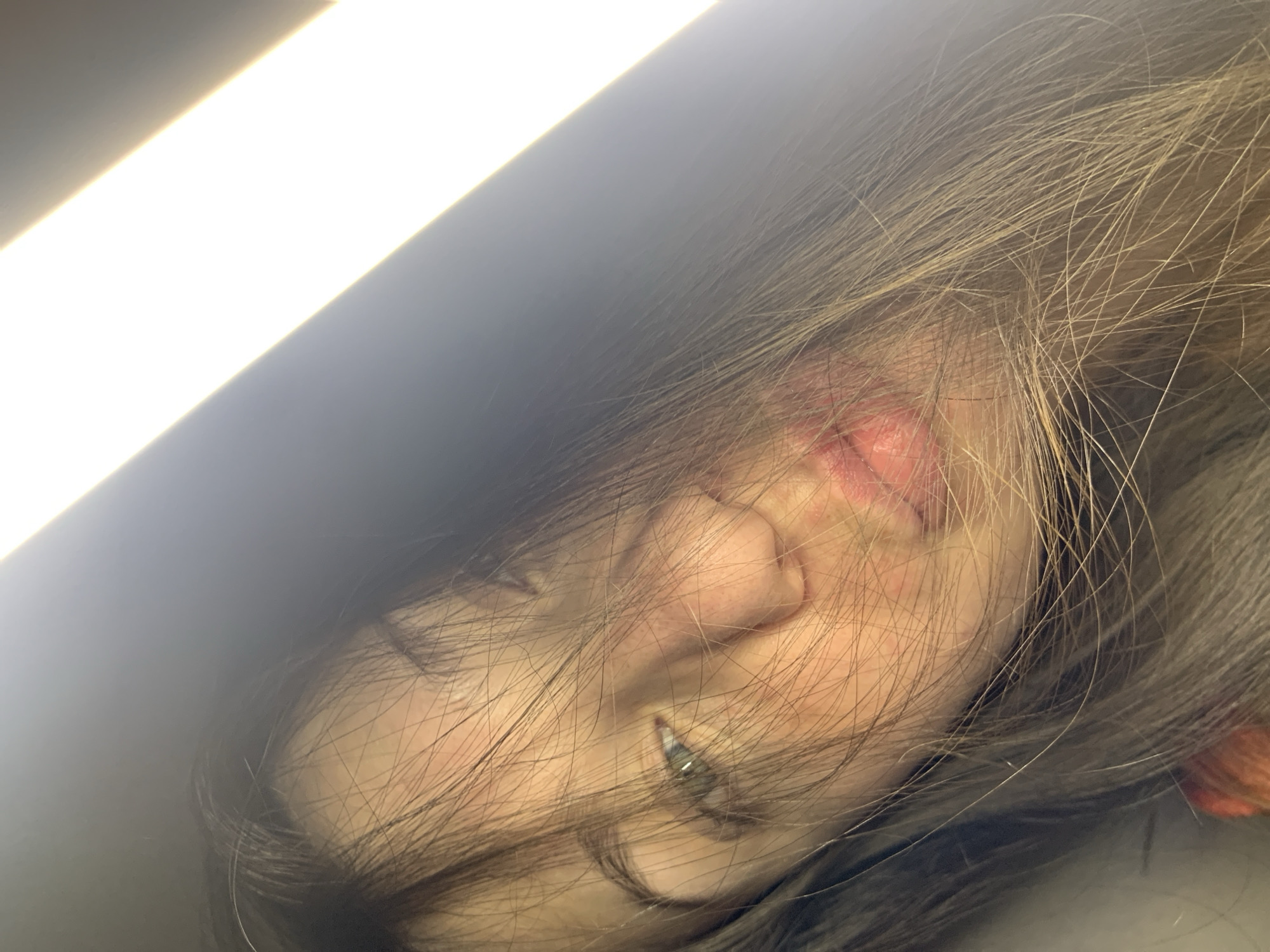 A close-up, tilted selfie of a young woman's face is captured against a bright light source. Her hair, a blend of brown and blonde, is dishevelled and partially obscures her features. One eye, a light green or hazel, is visible, looking slightly off-camera. Her lips are slightly parted and appear to have a rosy tint. The lighting creates strong highlights and shadows across her face, emphasizing the texture of her skin and hair. The background is dark, with a prominent diagonal band of bright white light. The location in Chaikovsky, Russia is not determinable from the image.