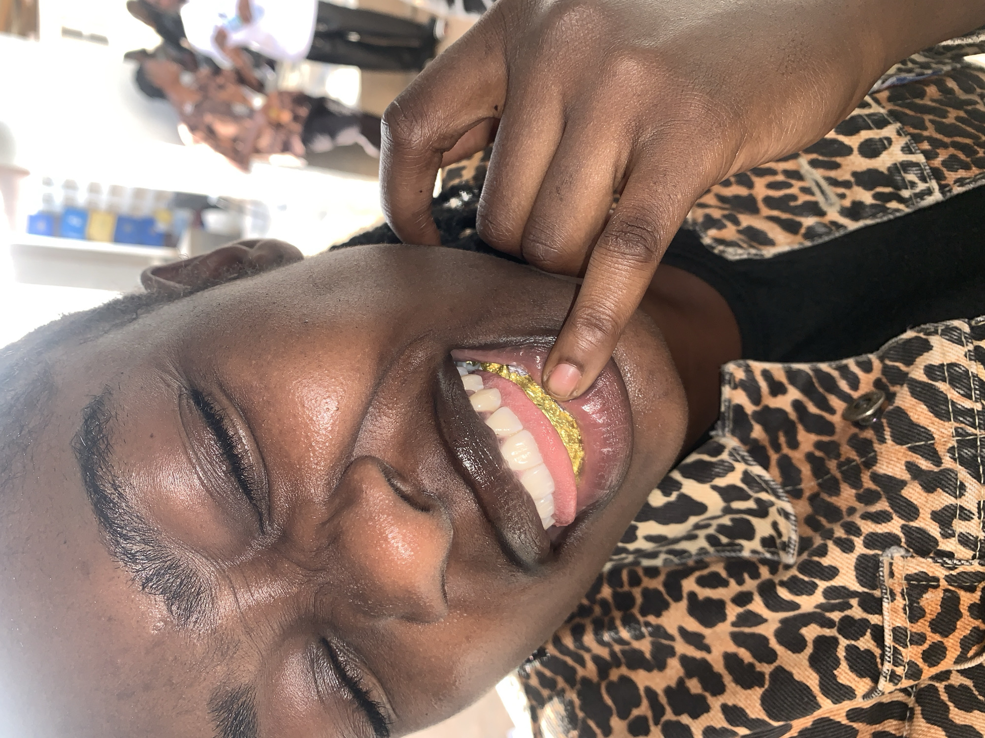 A close-up portrait depicts an individual with dark skin, eyes closed, and an expression indicative of amusement or joy. The subject is wearing a leopard print jacket over a black garment. The left hand, adorned with short, neat fingernails, is used to pull down the lower lip, exposing the dental arrangement. The upper teeth are visible, appearing white and evenly aligned. The lower teeth are covered by a prominent gold-colored dental grill, which extends over the gums and has a textured, possibly crinkled appearance. The skin exhibits a smooth texture with visible pores and natural highlights. In the blurred background, several indistinct figures are visible, one wearing a patterned shirt and another a white shirt. Further back, stacked blue and yellow containers are partially discernible against a light-colored wall. The scene is illuminated by bright, possibly natural light. This event is observed in the city of New Stadium, Nigeria.