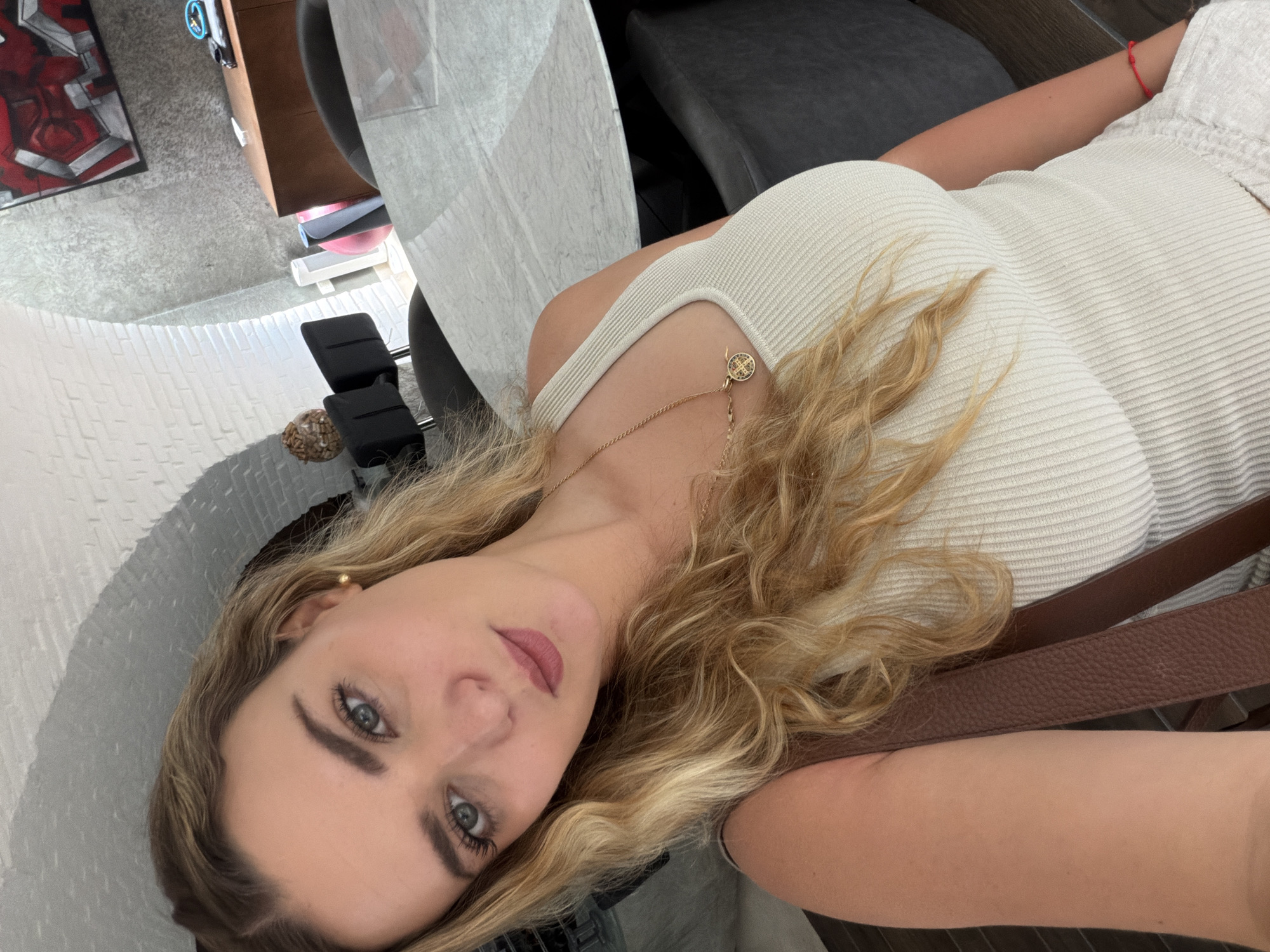 An angled, close-up indoor selfie shows a young woman with long, wavy blonde hair and blue-green eyes, looking directly at the camera. She has a calm expression and is wearing a light cream or off-white ribbed tank top. A gold chain necklace with a small, intricate round pendant adorns her neck, and a simple red string bracelet is visible on her left wrist. She wears a small gold stud earring in her right ear. Her makeup includes mascara, eyeliner, and a rosy-mauve lipstick.
The background, largely out of focus, reveals an indoor setting in San Bernardino Tlaxcalancingo, Mexico. On the left, a grey textured surface and a white, possibly brick-textured, curved wall are visible. A modern, red and grey geometric artwork hangs on a wall in the upper left corner. Dark furniture, including black padded chairs, is scattered in the mid-ground, suggesting a cafe, restaurant, or lounge environment. A small, dark, textured spherical object, resembling a chocolate, rests on a surface in the lower-left background. A brown leather strap, possibly from a bag or seat, crosses the lower right of the image. The lighting indicates it is daytime. No visible text is present.