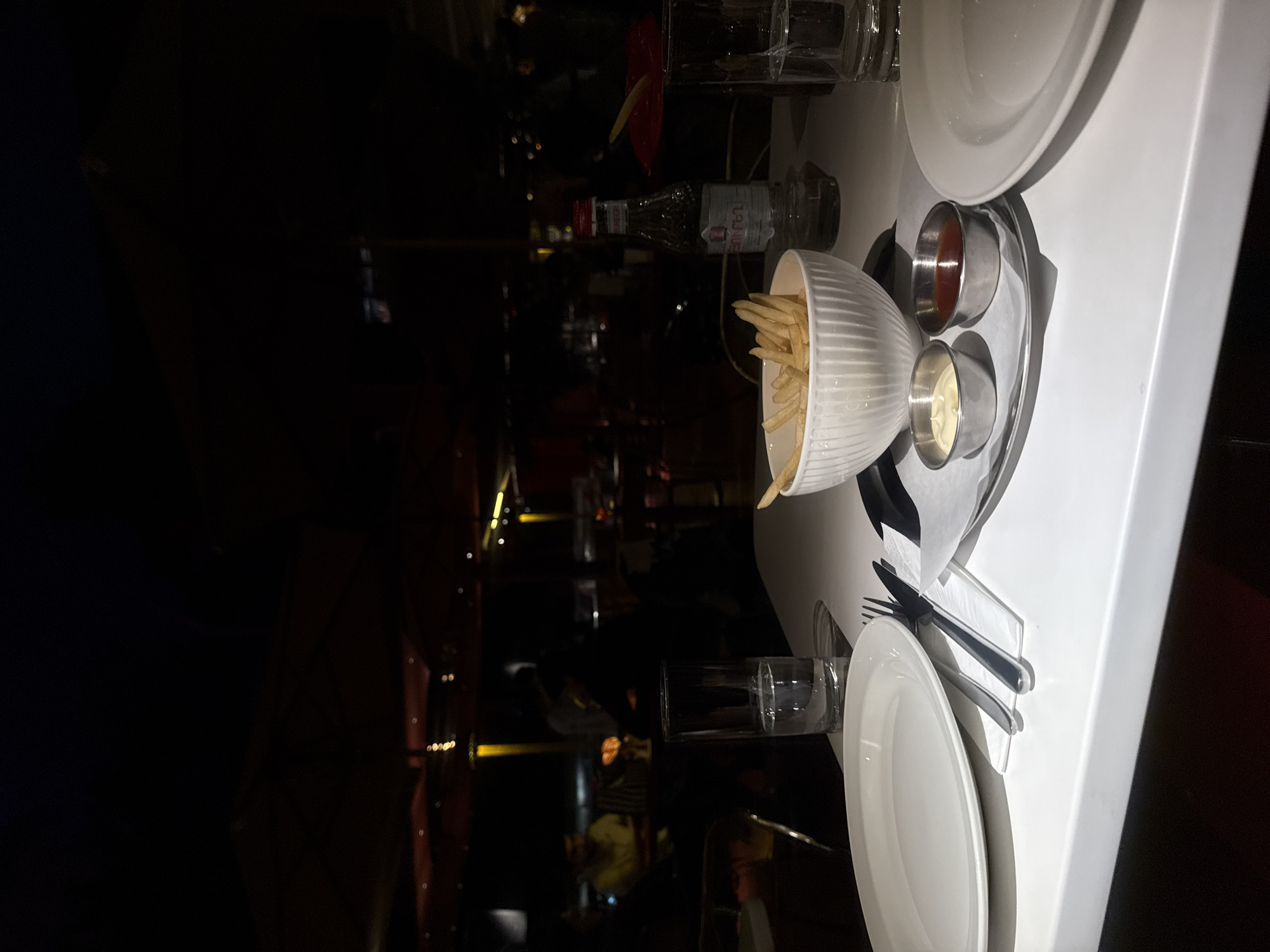 The image captures a dimly lit dining table in what appears to be a restaurant setting. The table, covered with a white surface, holds various items including plates, cutlery, a bowl of french fries, and small containers of dipping sauces (ketchup and mayonnaise). A miniature bottle of Taj Vue vodka is visible, along with glasses, one containing a decorative red flower. The background is dark, suggestive of a low-light environment within the restaurant. Given the presence of Taj Vue vodka and the provided location information, it is highly probable that this scene is located in Yerevan, Armenia. There is no explicit evidence of people in the image, but it can be inferred that the table is set for diners. The overall impression is of a meal being served or enjoyed in a dimly lit restaurant.