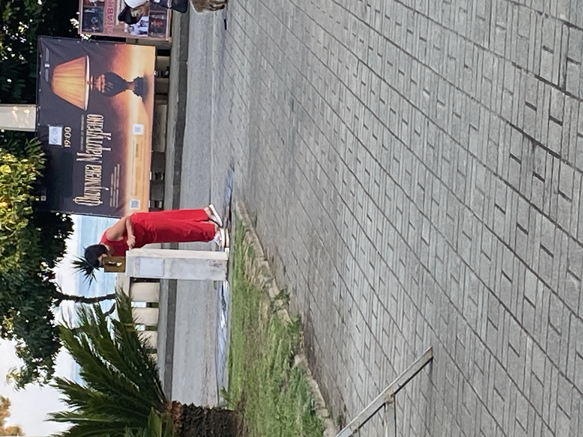 A woman in a red dress is leaning over a white concrete structure, possibly a fountain or a monument. She is looking down at something on the structure, her hands are resting on it. A palm tree is visible to her left, and a large banner with an image of a lamp and text in Cyrillic is behind her. The banner appears to be an advertisement for an event titled "Филумена Мартурано" with a time of 19:00. In the background, trees and the sea are visible. The scene appears to be outdoors, on a paved area with a pattern of rectangular bricks. A portion of a metal structure, possibly a chair or stand, is in the lower left corner. A dog is visible to the right, partially obscured. The setting is likely a public space in the city of Sokhumi, Abkhazia.