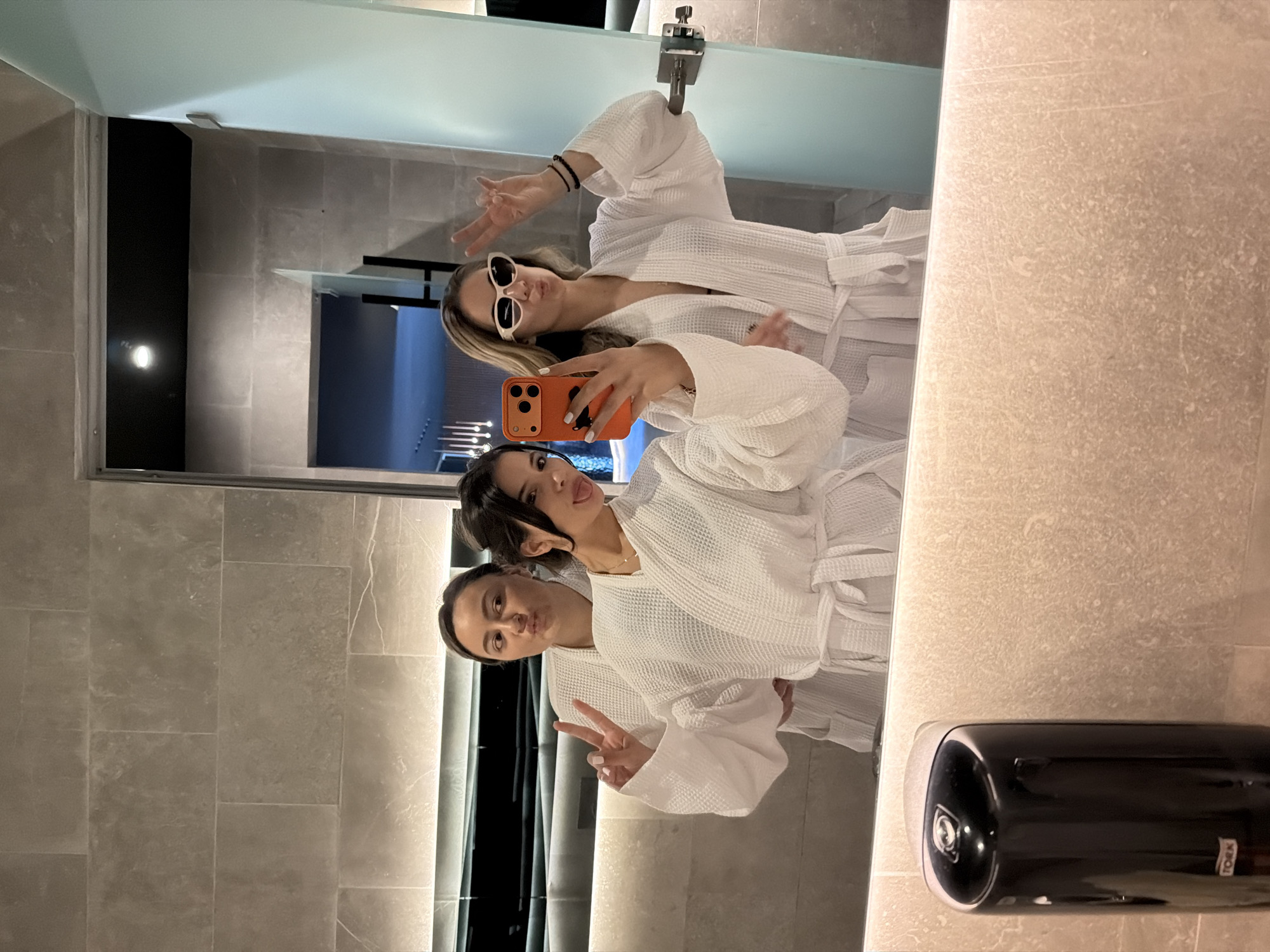Two women in white bathrobes are taking a selfie in front of a mirror. One woman is holding an orange phone. The location appears to be a spa or bathroom in Bania, Bulgaria. The background shows a glimpse of a pool or water feature through an opening. There is a black soap dispenser in the foreground. The women are posing and making gestures.