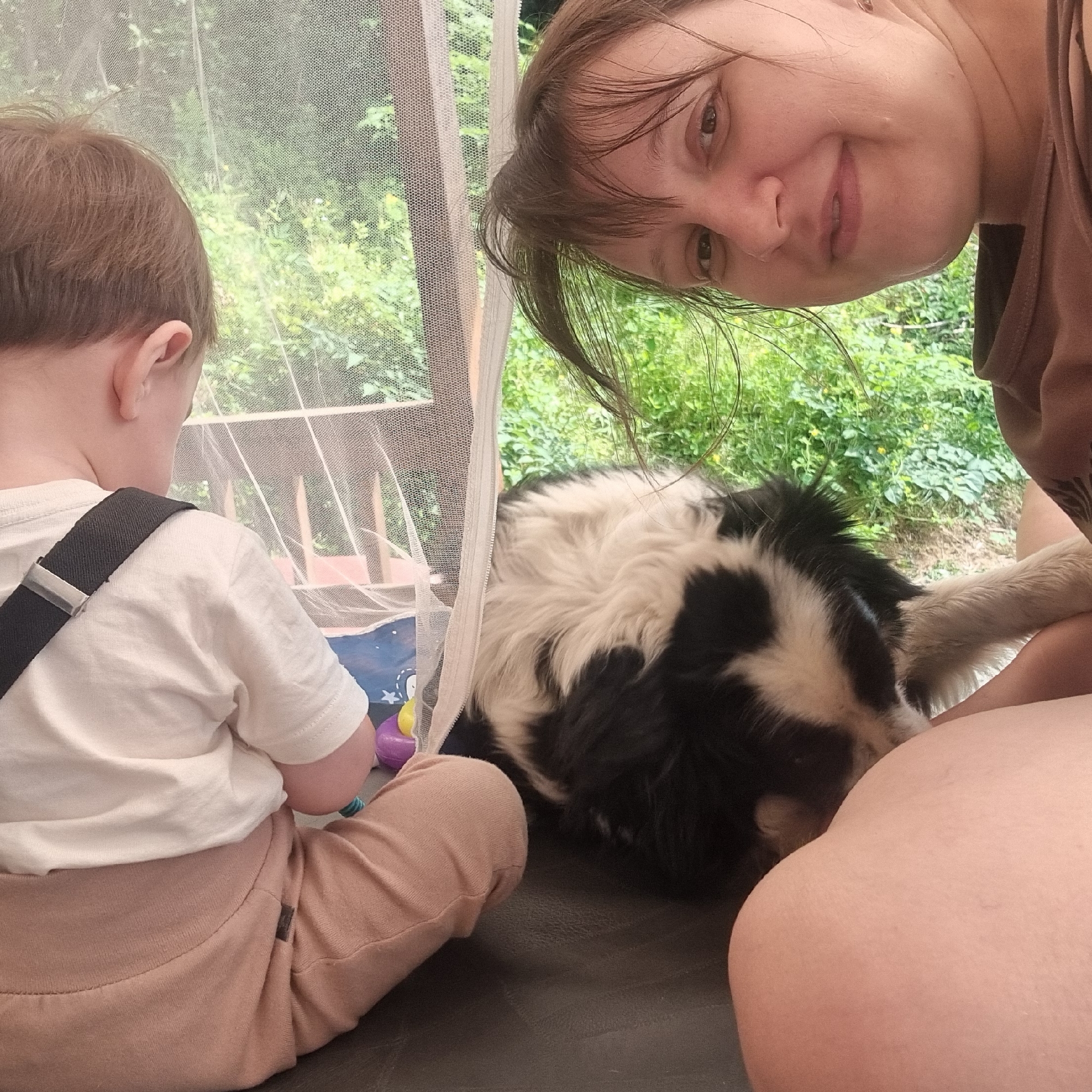 In an outdoor setting, likely Dunnae-myeon, South Korea, a woman is taking a selfie with a child and a dog. The woman's face is partially visible, looking towards the camera with a slight smile. The child, from behind, is wearing a white t-shirt with suspenders and light brown pants, and appears to be interacting with the dog. The dog, a black and white breed, is also partially visible and seems to be close to the child. The background consists of lush green foliage, suggesting a natural or rural environment. A sheer mesh curtain is visible to the left, possibly part of a tent or outdoor shelter, separating the subjects from the outside environment. The overall scene suggests a casual, familial moment captured outdoors.