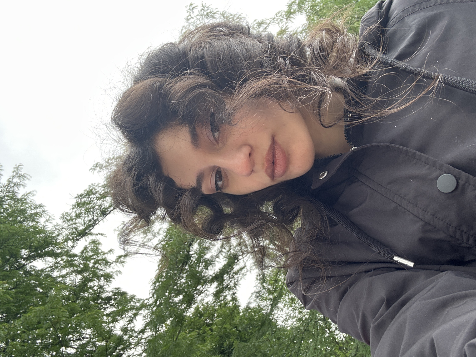 A close-up, low-angle, outdoor, selfie-style photograph depicts a young woman with dark, curly hair. She is wearing a black jacket. Her head is tilted slightly back, and her gaze is directed towards the camera. Green trees and a cloudy sky are visible in the background. The image does not provide any specific information about the location in the city of Sofia, Bulgaria.