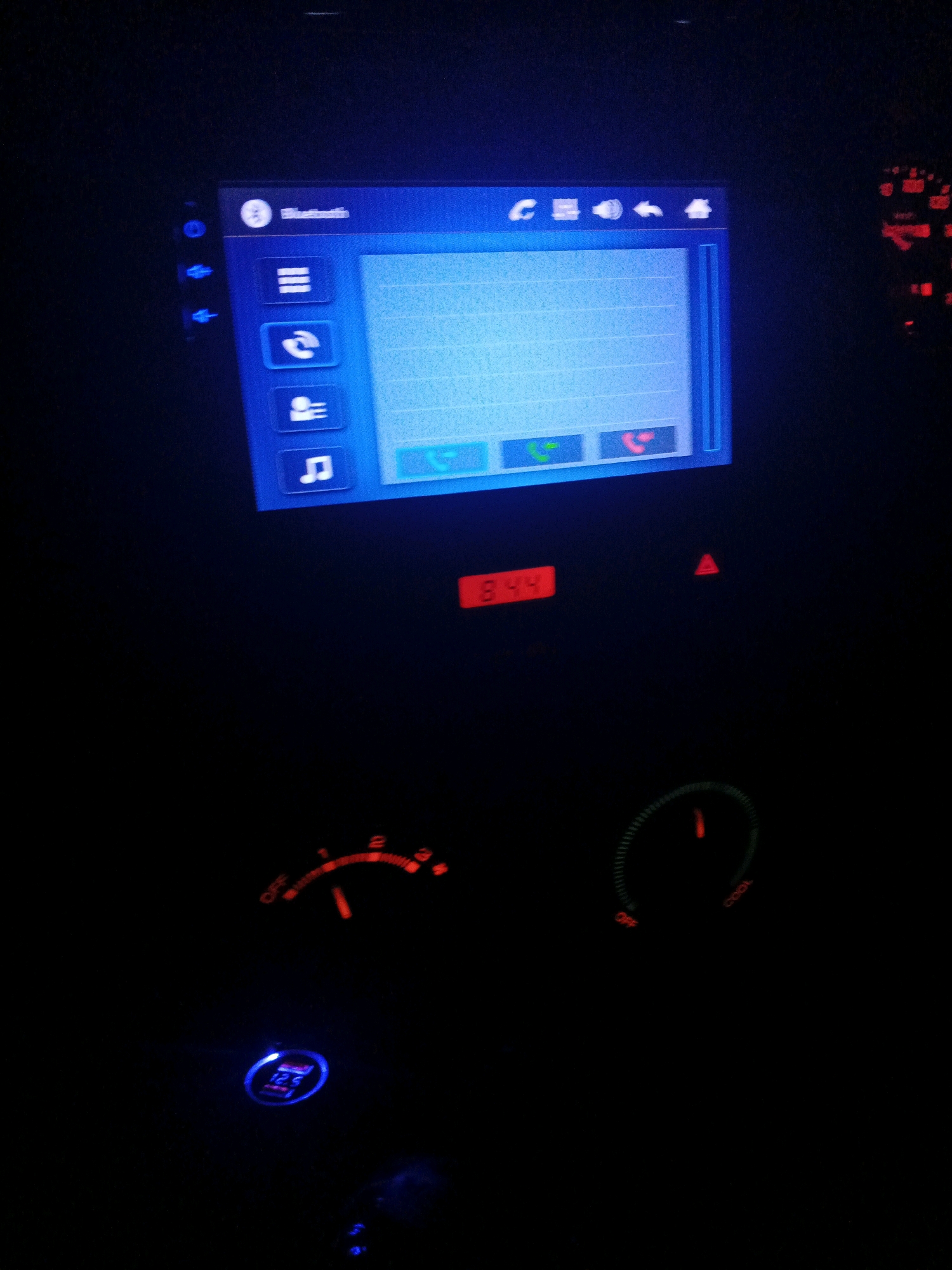 The image displays the interior of a vehicle at night, possibly in Sidoharum, Indonesia, showcasing the dashboard and a car stereo system. The car stereo, illuminated in blue, features a screen displaying a list of options, including "Bluetooth." Below the screen, there are icons for various functions. To the right of the stereo, a portion of a car's speedometer is visible, with red illuminated needles indicating speed. Below the stereo, a red digital display shows "8:44". Further down, two circular gauges are present: one with a red-glowing needle and markings labeled "OFF" and numbers, and another with a green-glowing needle and markings like "OFF" and "COOL". A USB charging port with a blue light is also visible. The overall scene is dark, emphasizing the illuminated elements of the dashboard.